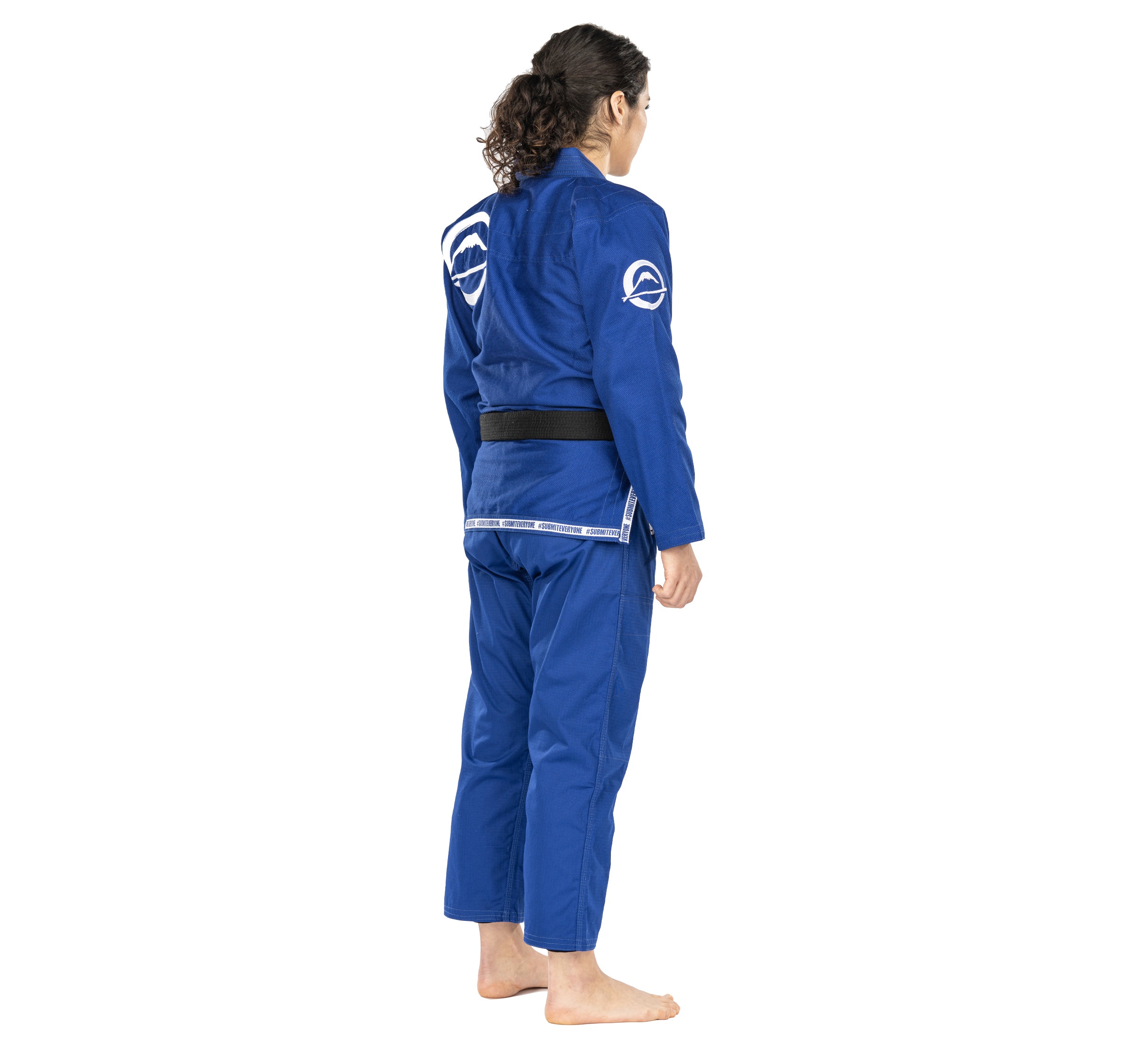 A woman with long hair in a ponytail wears the Submit Everyone Womens BJJ Gi Blue and a black belt, standing barefoot and facing away from the camera against a white background.