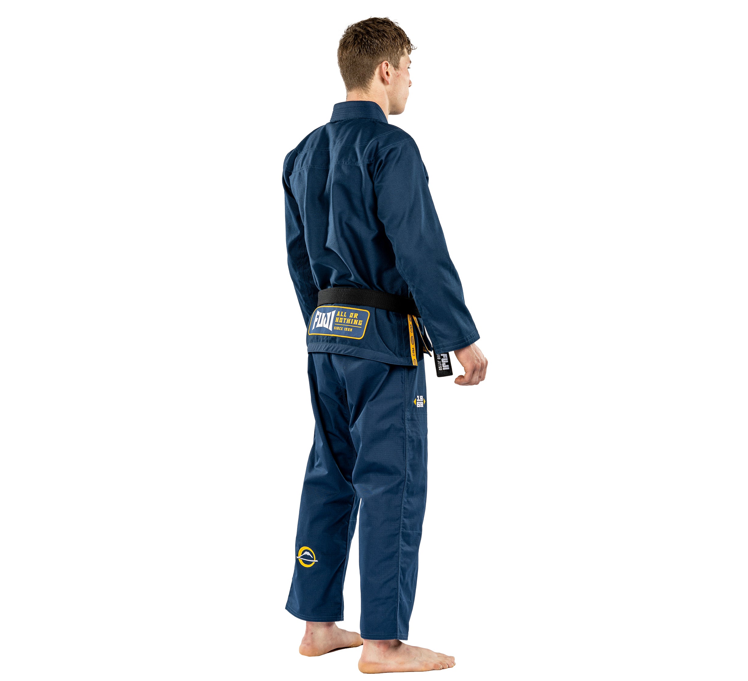 A person stands barefoot, facing away from the camera, wearing the Suparaito BJJ Gi Worldwide Edition Navy with patches and a black belt against a white background.