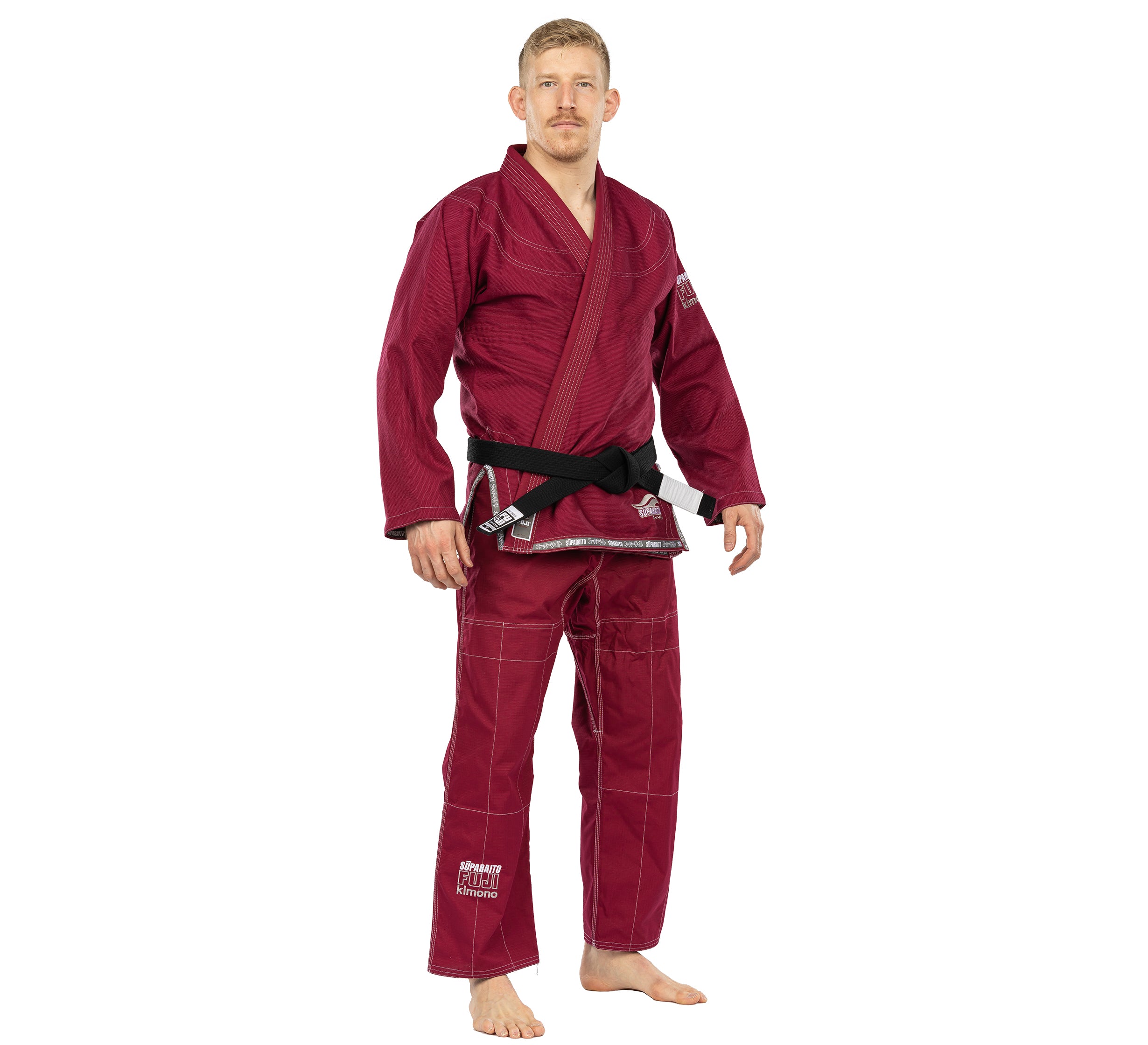 A man with short blond hair and a mustache wears a Suparaito BJJ Gi in burgundy with a black belt. He stands barefoot, facing forward with a neutral expression against a plain white background.