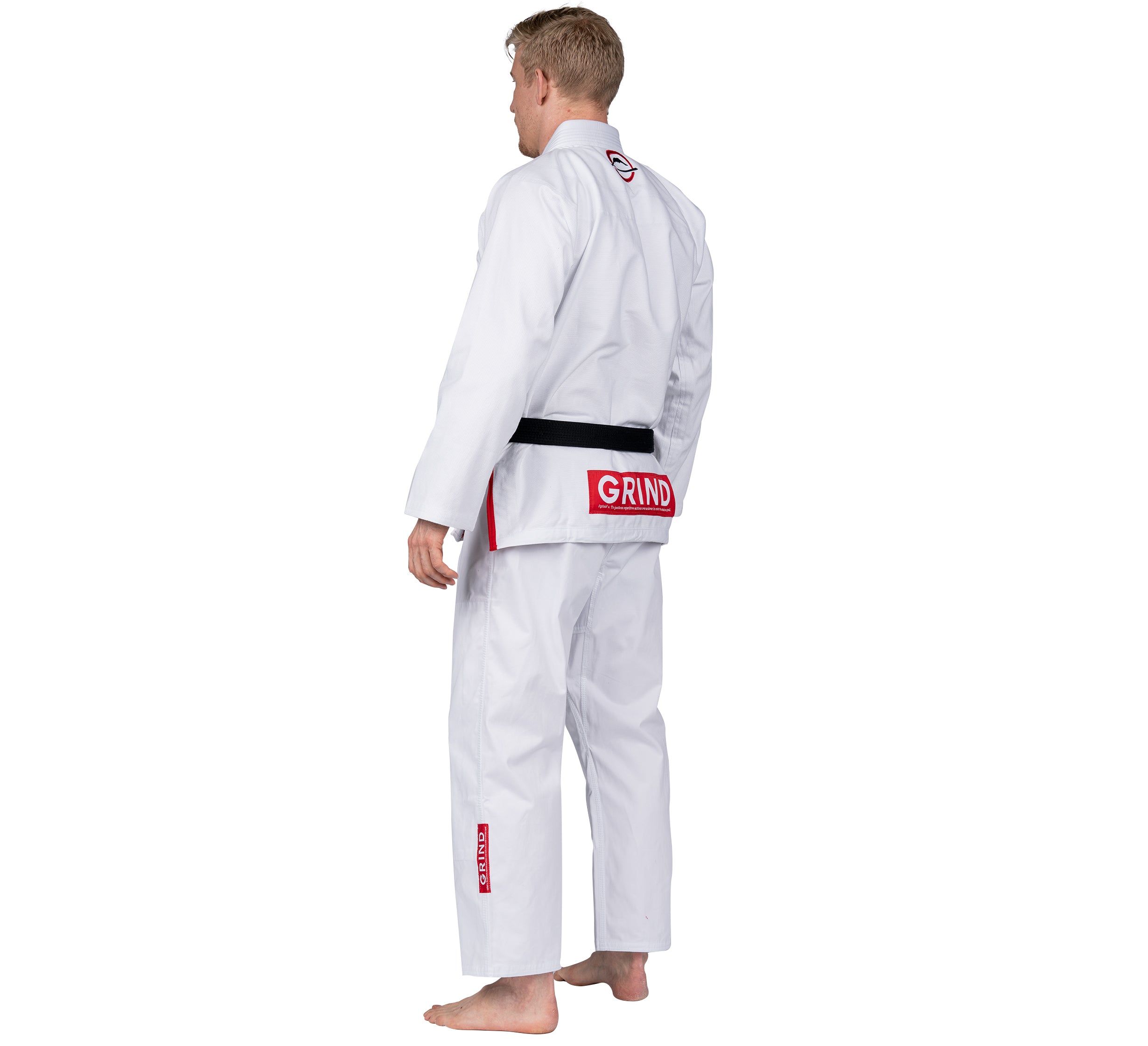 A barefoot person stands facing away, wearing the Elemental BJJ Gi Grind White with a black belt. The white gi features red patches, including one above the belt that says GRIND.