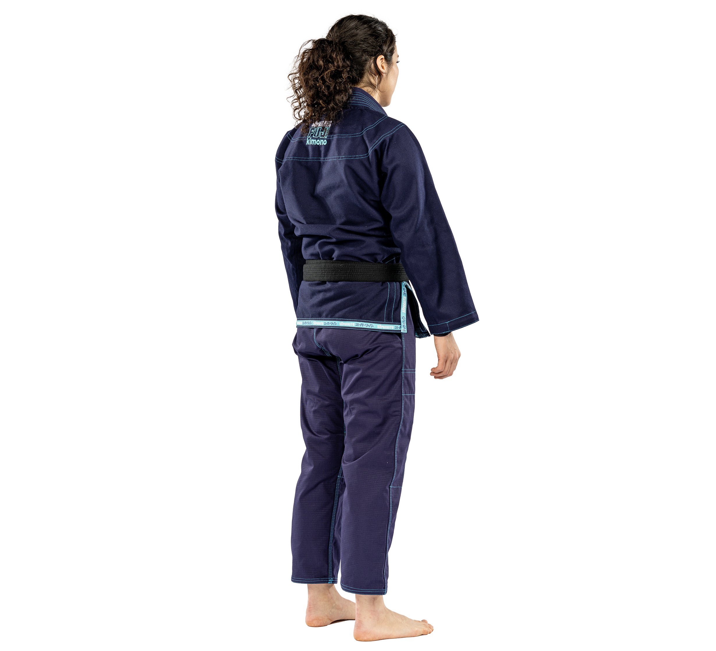A person with long curly hair wears the Suparaito Womens BJJ Gi in navy and a black belt, standing barefoot and facing away from the camera against a white backdrop.