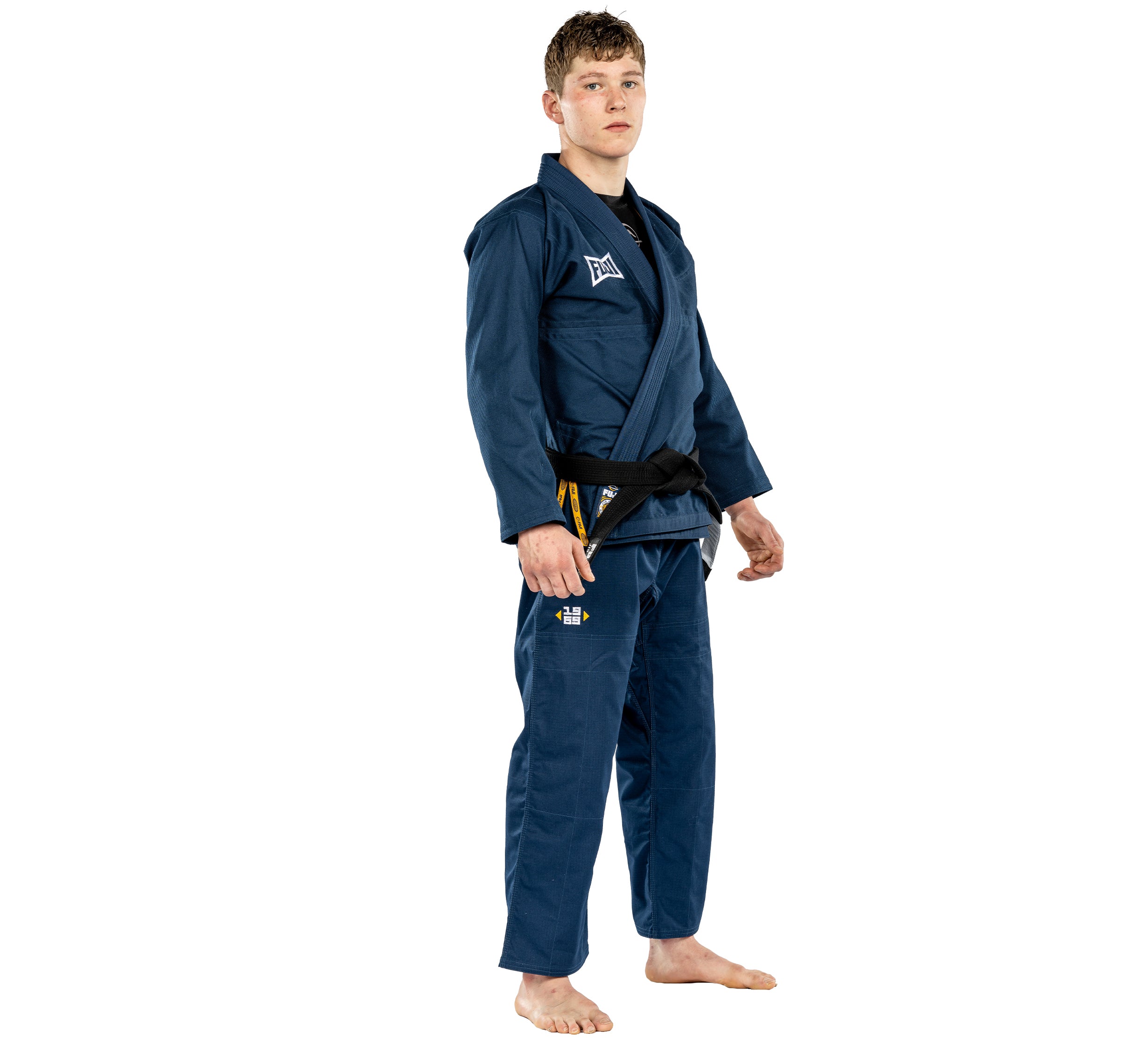 A young person wears the Suparaito BJJ Gi Worldwide Edition Navy with a black belt, standing barefoot against a white background and looking forward with a neutral expression.