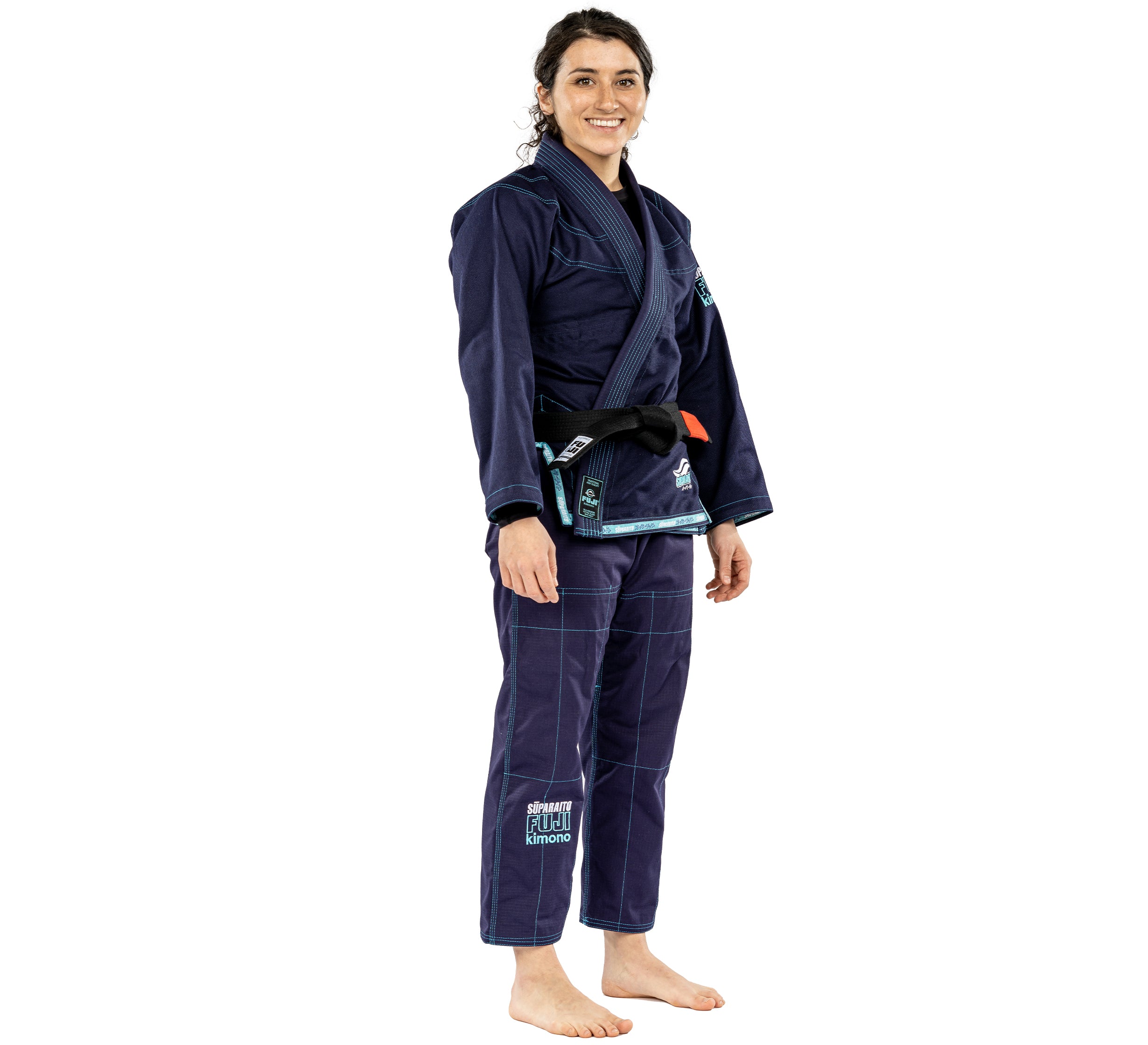 A woman smiles barefoot in the Suparaito Womens BJJ Gi Navy with a black belt. The gi features light blue stitching and patches, and she stands against a plain white background.