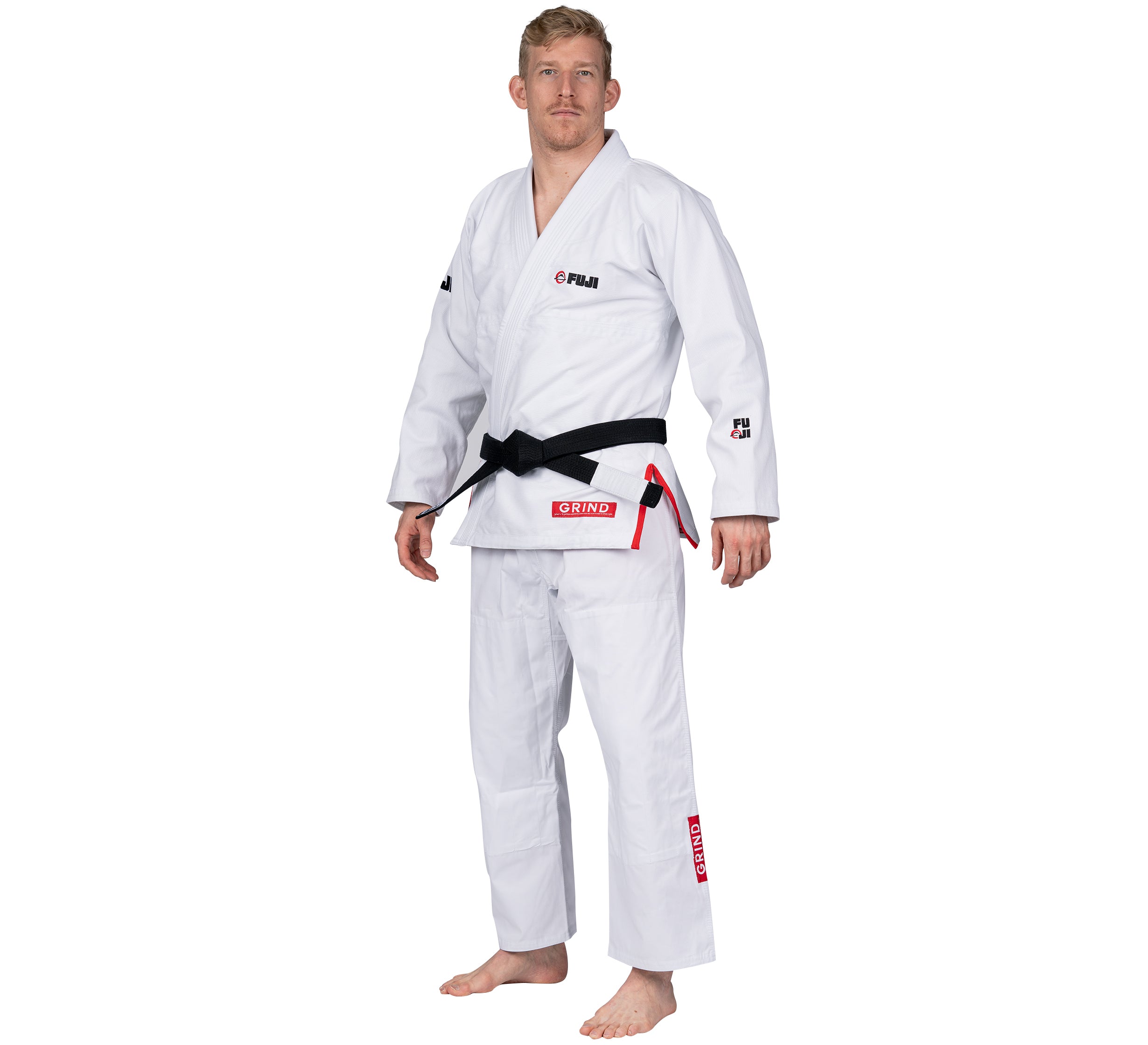 A man stands barefoot, wearing the Elemental BJJ Gi Grind White with a black belt. The gi features red and black embroidered patches and logos. He faces slightly to the side against a plain white background.