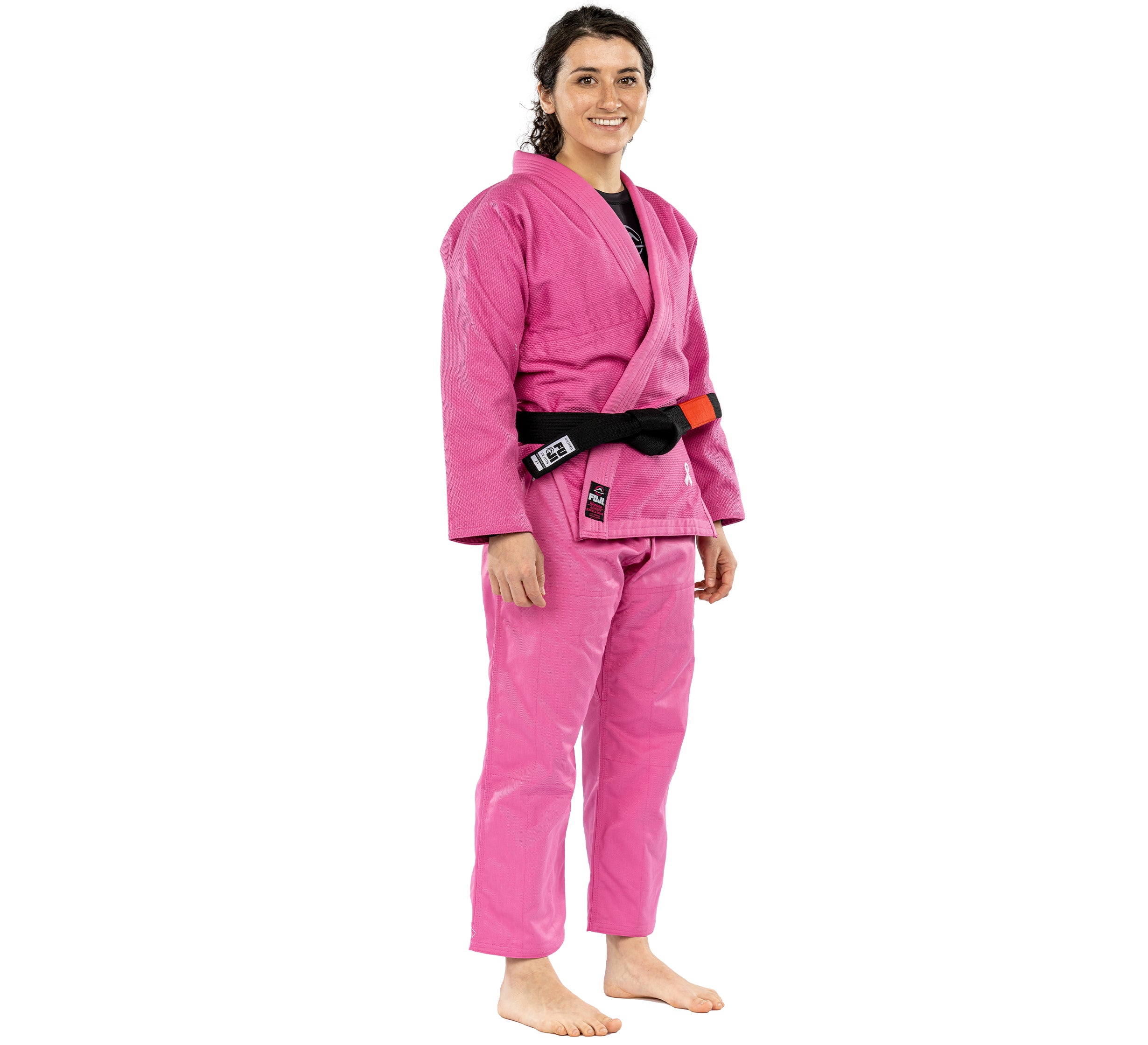 A woman wearing the All Around Womens BJJ Gi Pink with a black belt stands barefoot and smiling against a white background.