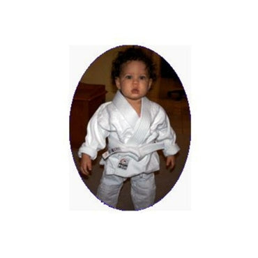 A young child with curly hair stands indoors, wearing a white FUJI Baby Judo Gi and white belt, looking at the camera.