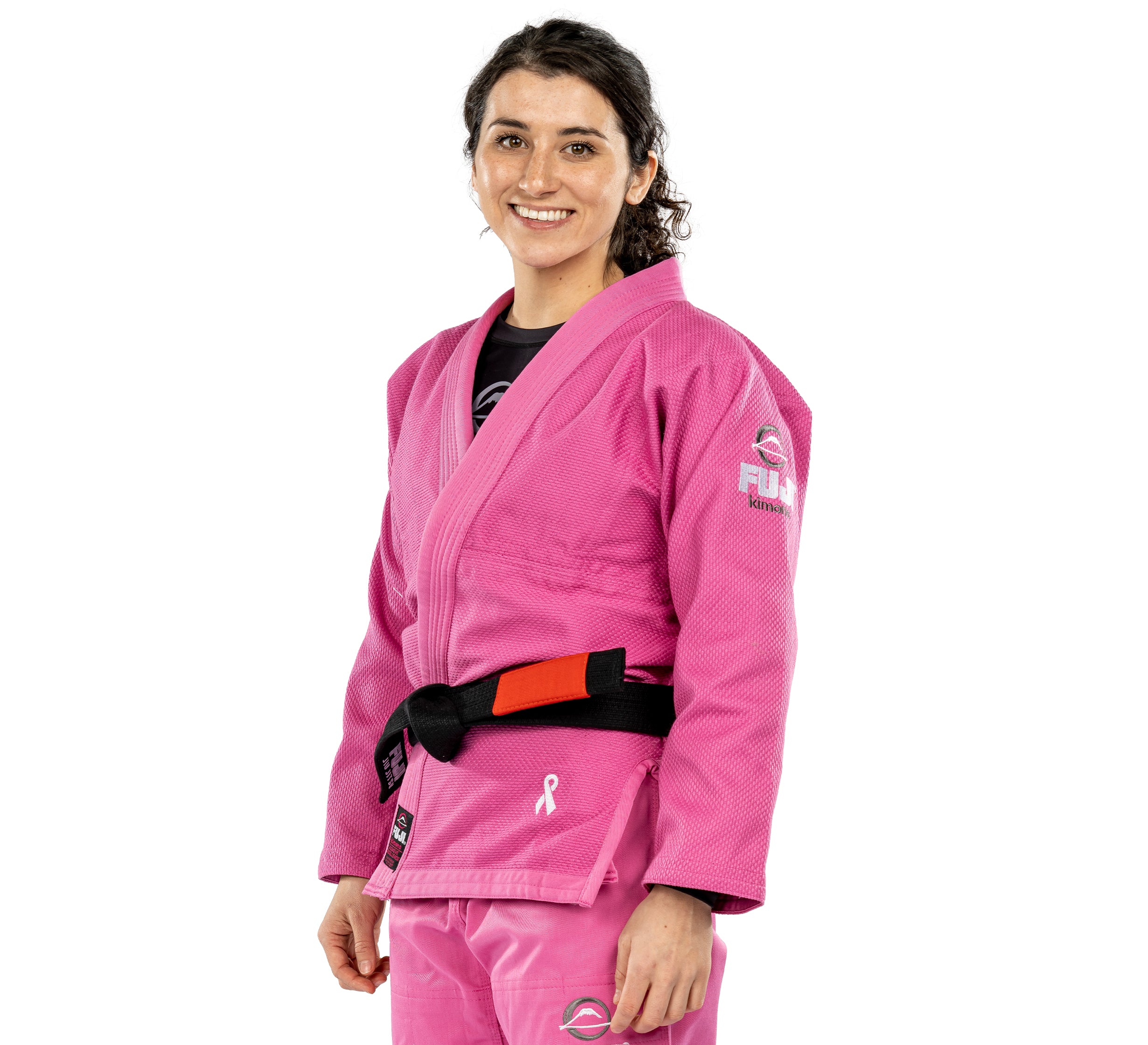 A woman smiles wearing the All Around Womens BJJ Gi Pink with a black belt featuring a red bar. The gi displays various patches, including a small pink ribbon on the lower part to symbolize breast cancer awareness.