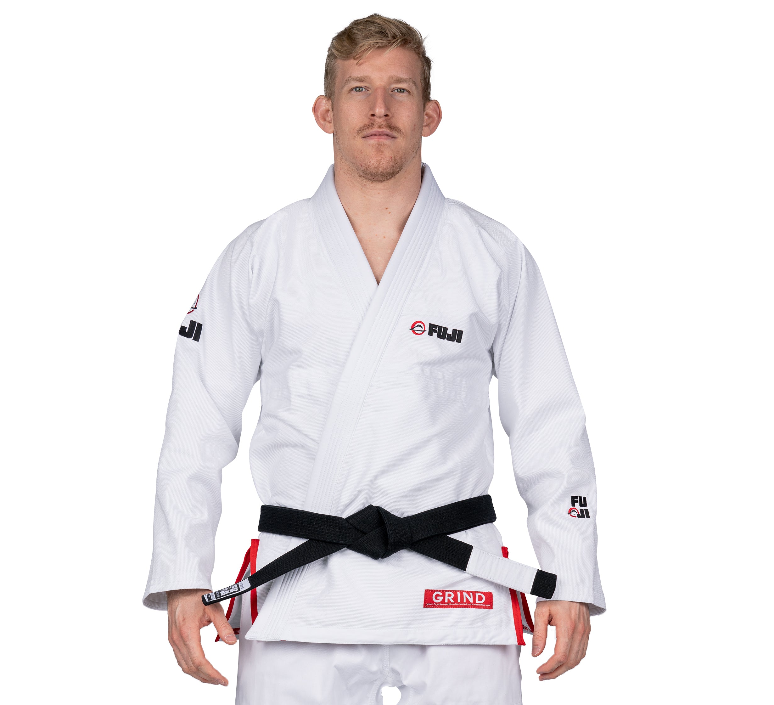 A man with short blonde hair and a neutral expression stands facing forward, wearing the Elemental BJJ Gi Grind White with FUJI logos on the chest and sleeves and a GRIND patch near the waist, paired with a black belt.