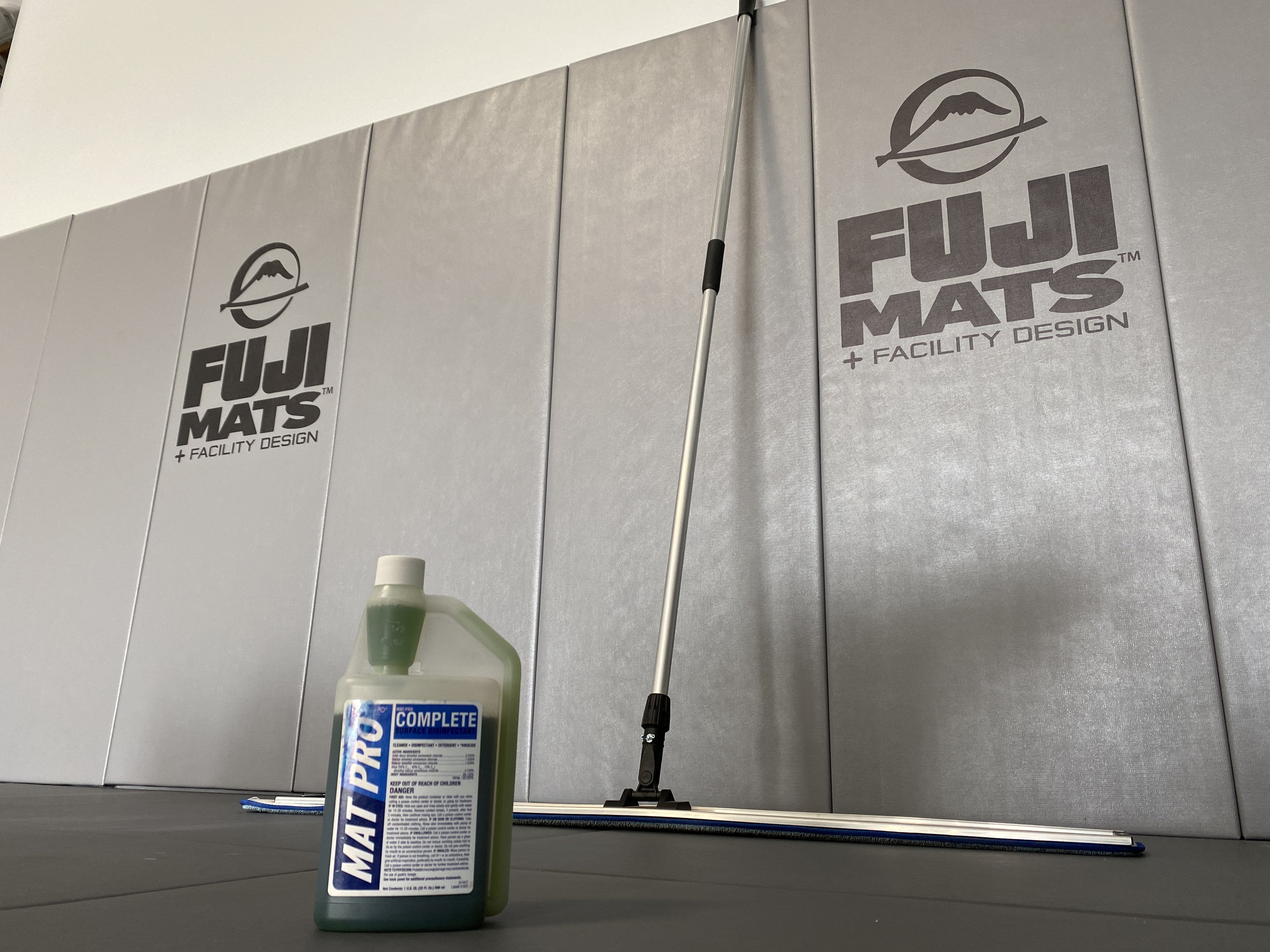 A bottle of EZ-Pour Concentrated Mat Cleaner 32 oz and a floor mop sit on black mats in front of a gray wall with FUJI MATS + FACILITY DESIGN printed on it.