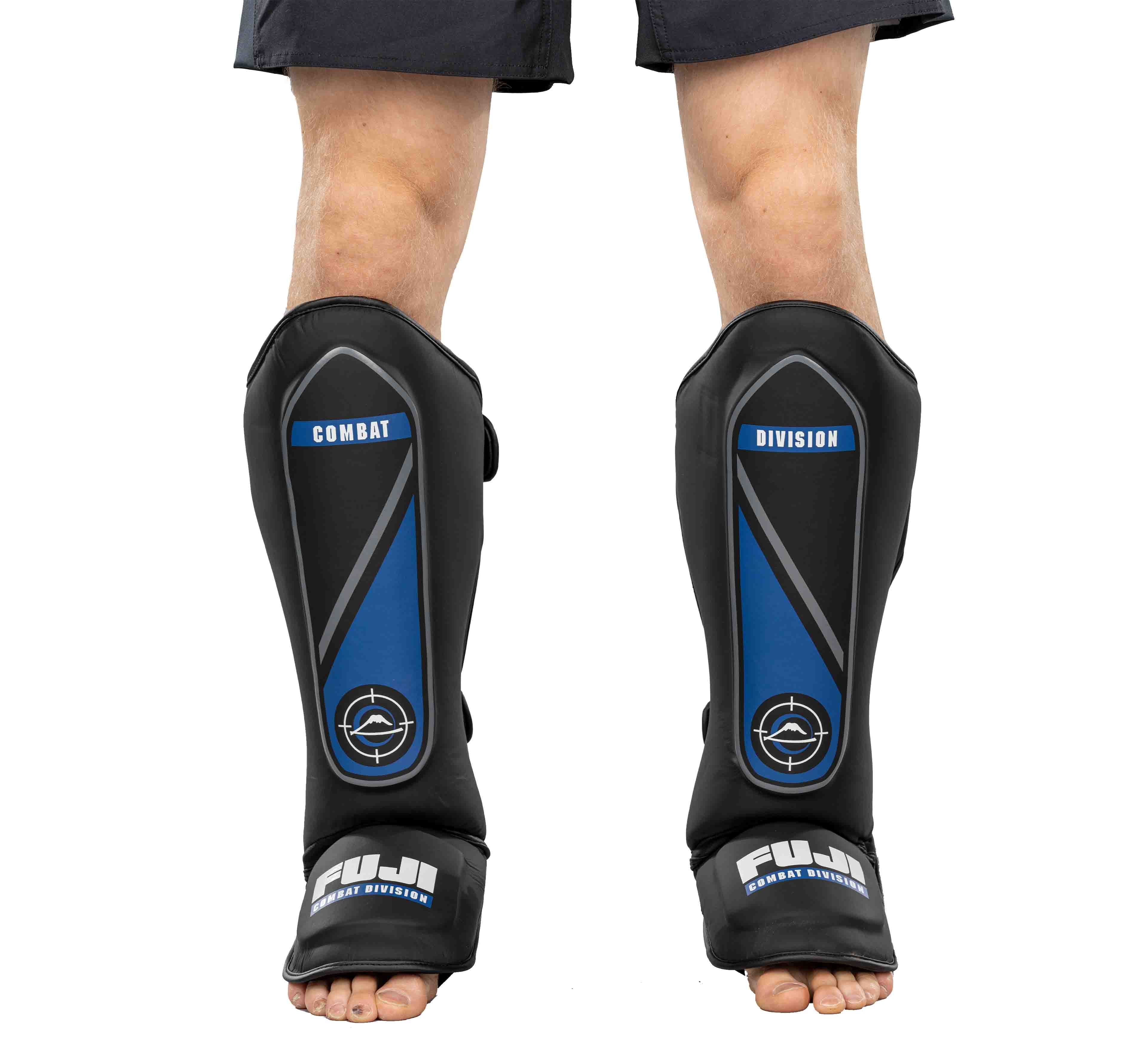 A person wears black and blue Precision Striking Shin Guards while standing barefoot, with black shorts visible above the knee.