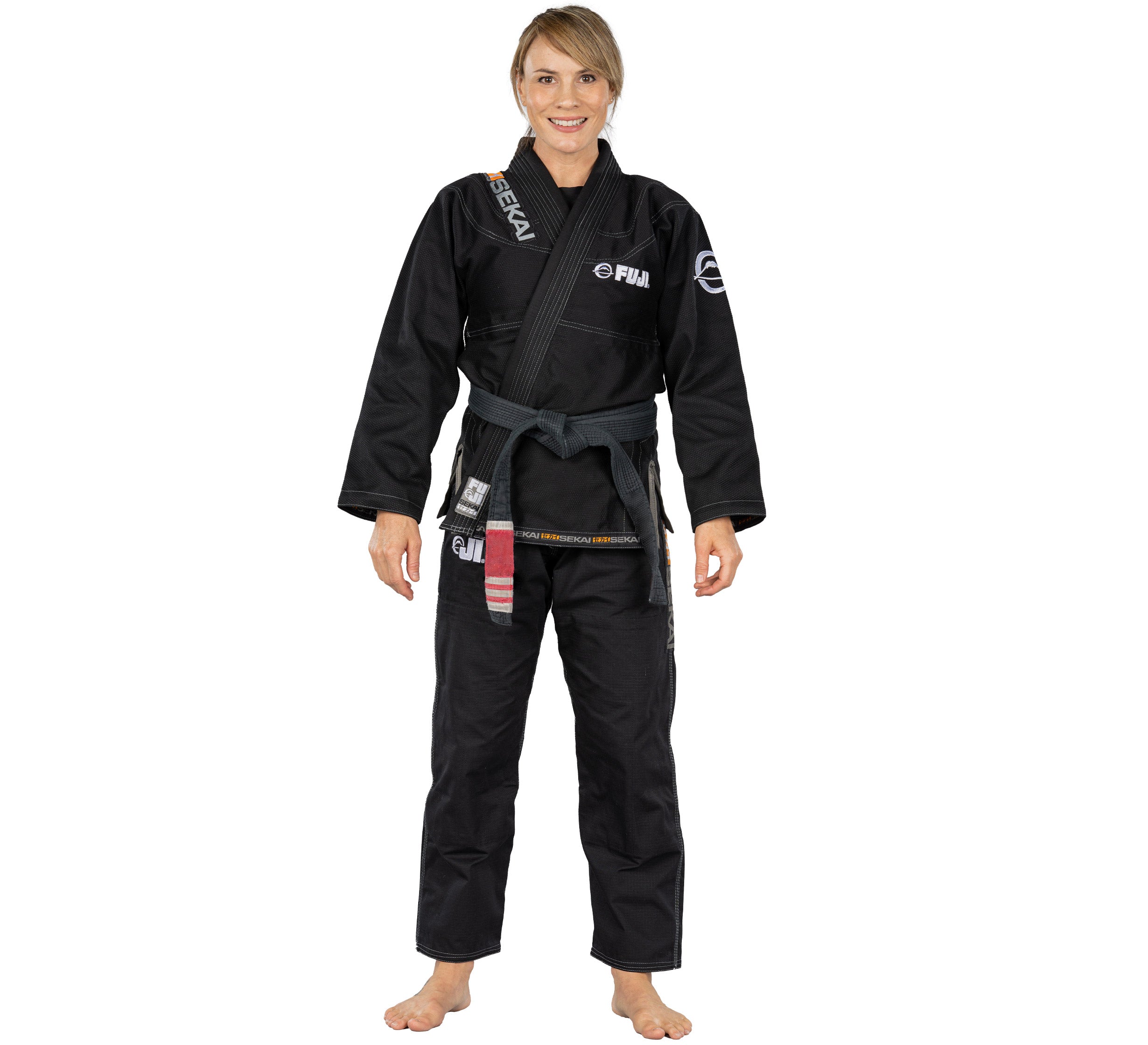A woman stands barefoot and smiles at the camera wearing the Sekai 2.0 Womens BJJ Gi Black with various patches and a black belt, against a plain white background.