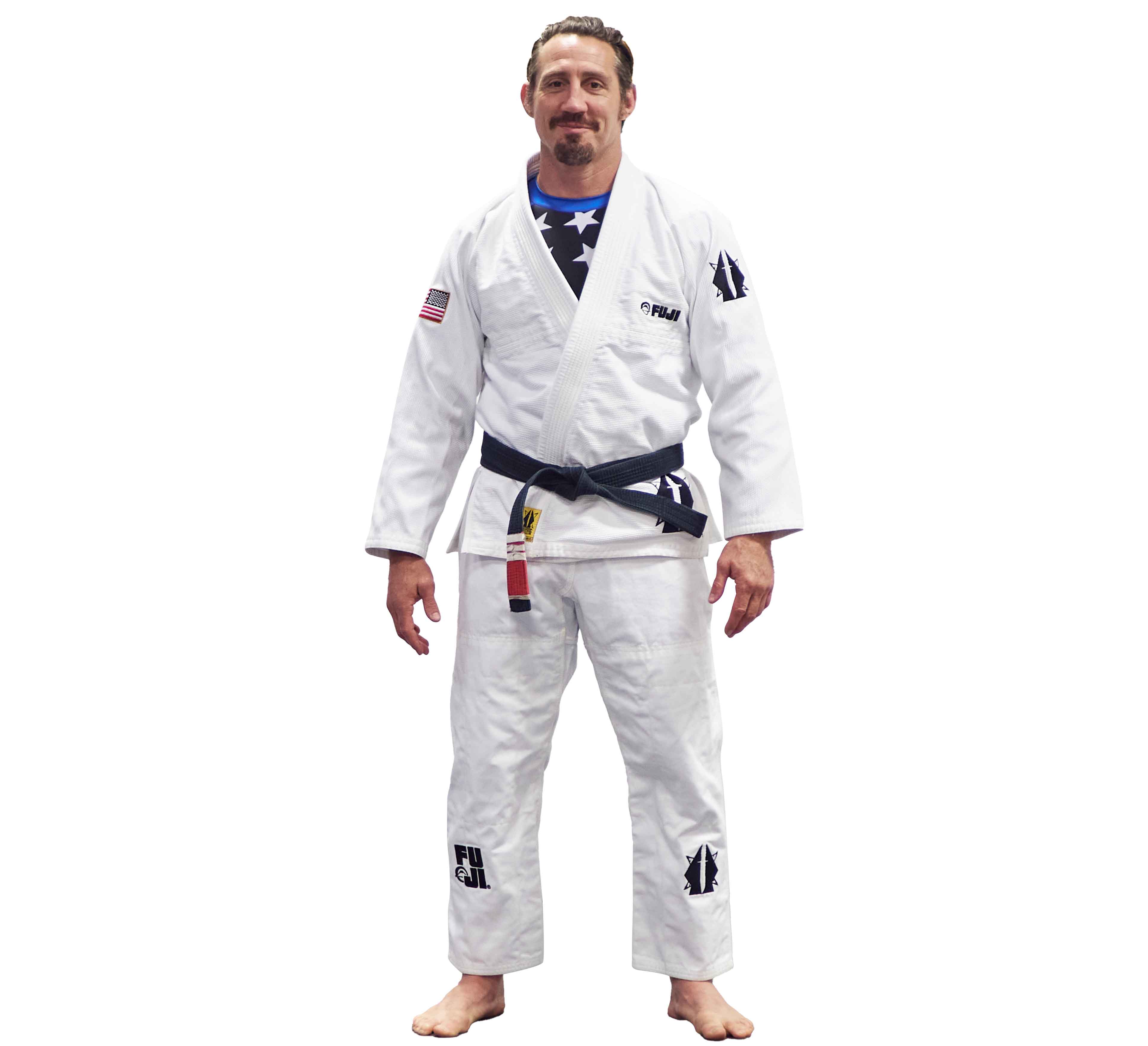 A man with short hair and a trimmed beard stands barefoot, smiling, in the Lightweight BJJ Gi Sheepdog Response Edition White, featuring patches and an American flag on the shoulder. He wears a black belt.