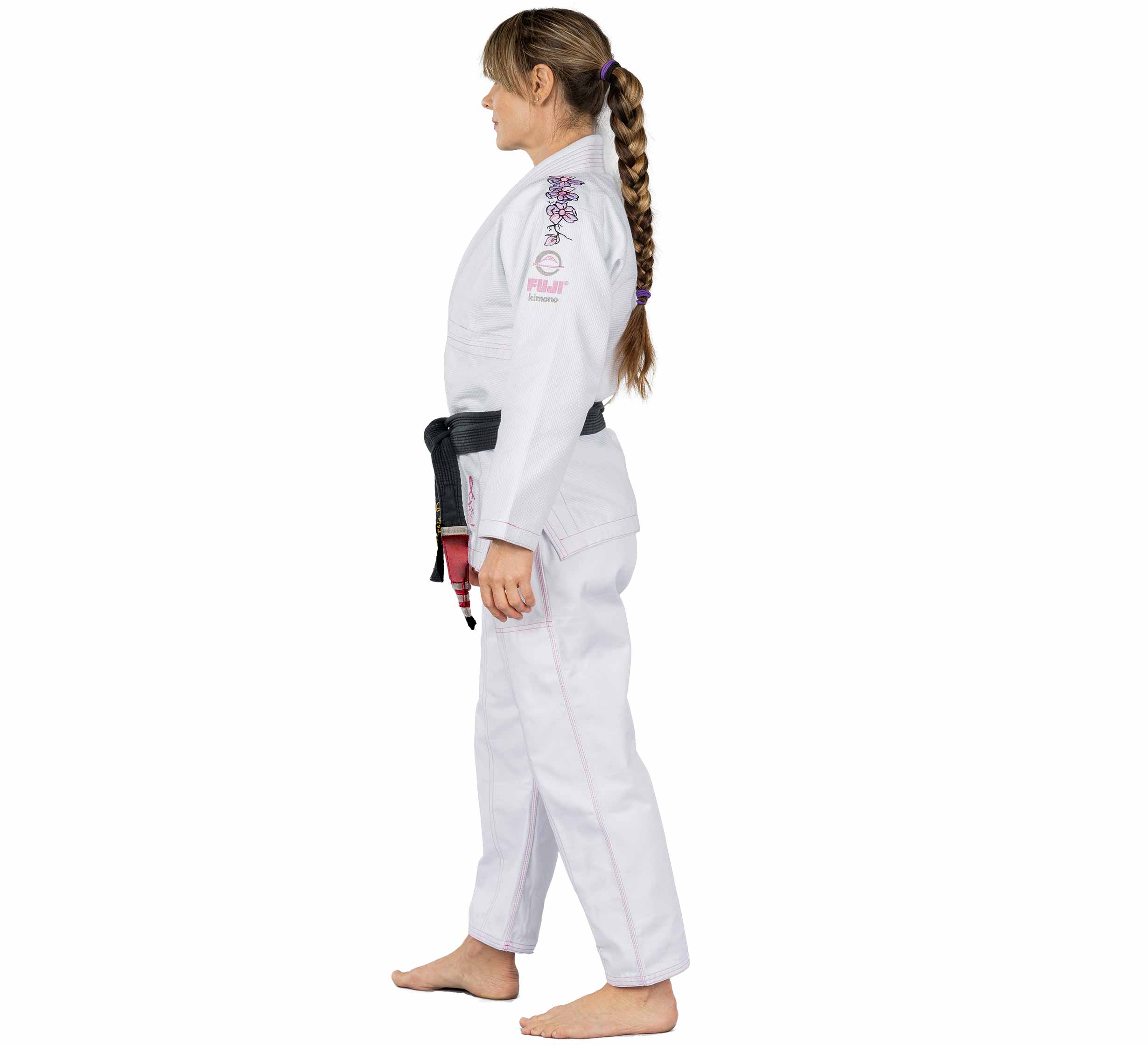 A barefoot woman with a long braid wears the Blossom Womens Jiu Jitsu Gi White, featuring pink trim and a black belt, as she faces left against a plain white background.