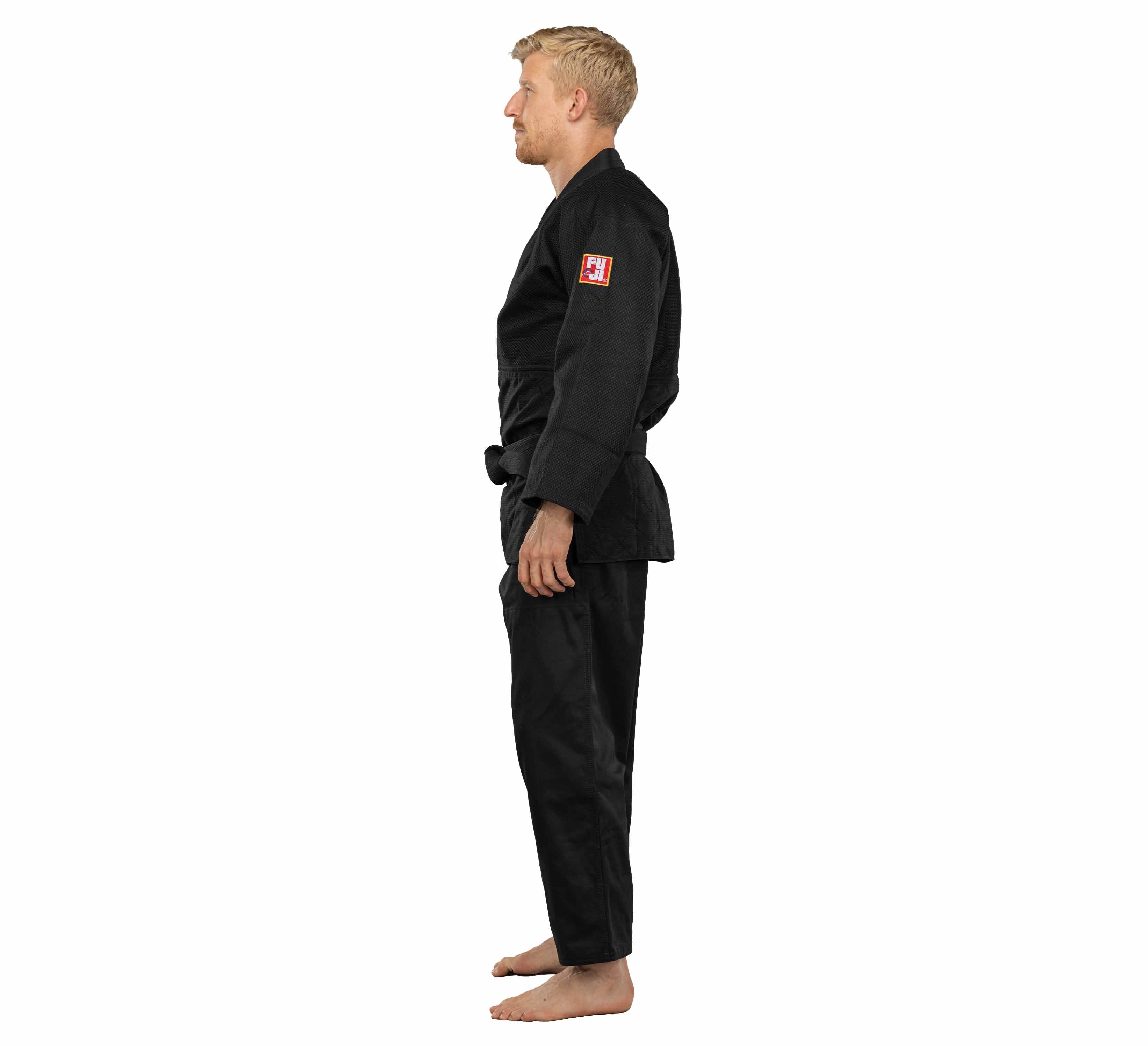 A barefoot man wearing the Euro Competition Judo Gi Black stands in profile against a white background, facing left with his arms relaxed at his sides.