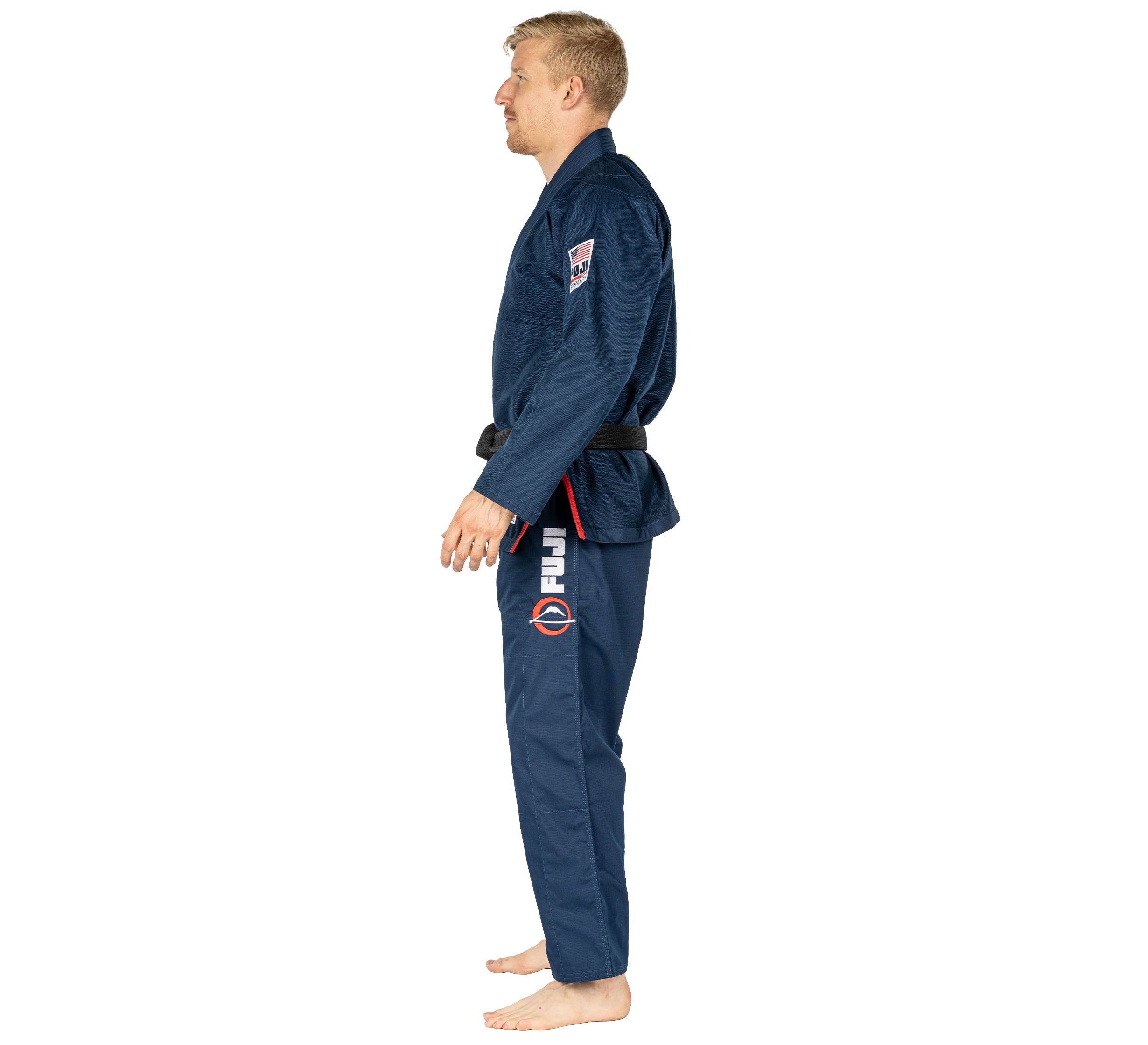 A man stands barefoot in profile, wearing the Suparaito BJJ Gi 1776 Americana Edition Navy with a black belt. The gi features patches and logos on the sleeve and pants, set against a plain white background.