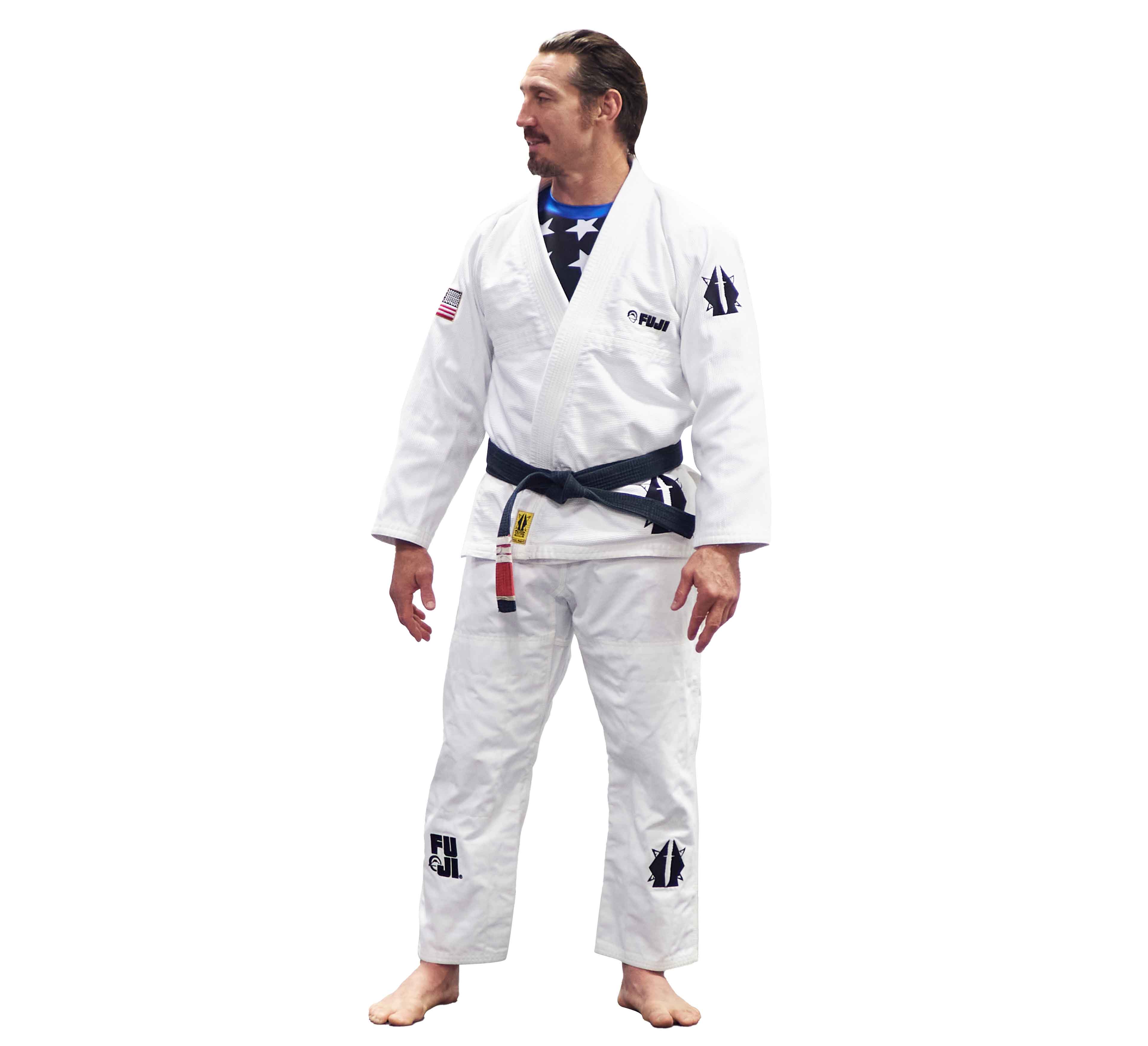 A man stands barefoot and looks left, wearing the Lightweight BJJ Gi Sheepdog Response Edition in white, featuring various patches and a black belt.