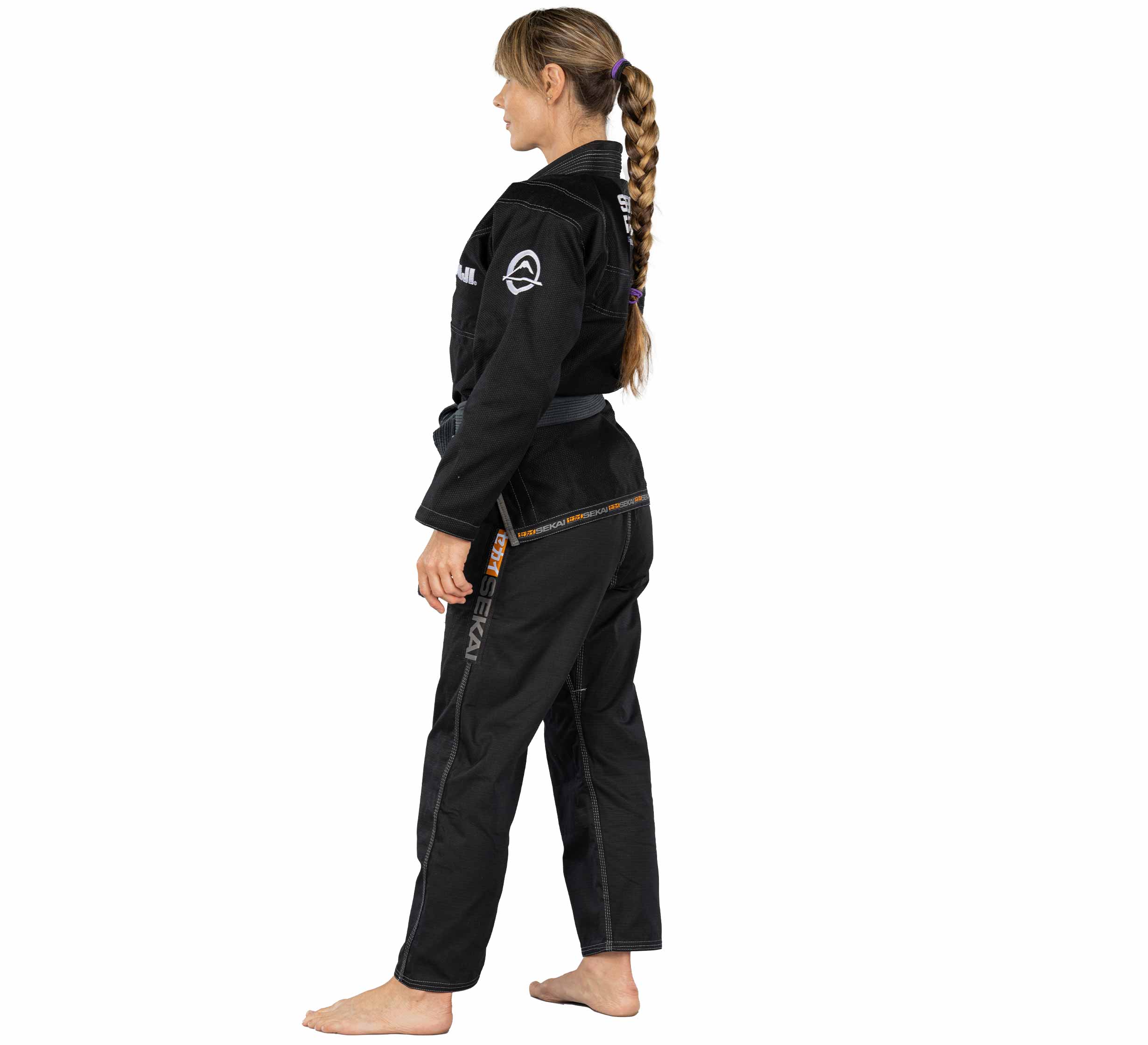 A woman with long braided hair stands barefoot in profile, wearing the Sekai 2.0 Womens BJJ Gi Black with a belt and patches on the pants, against a plain white background.