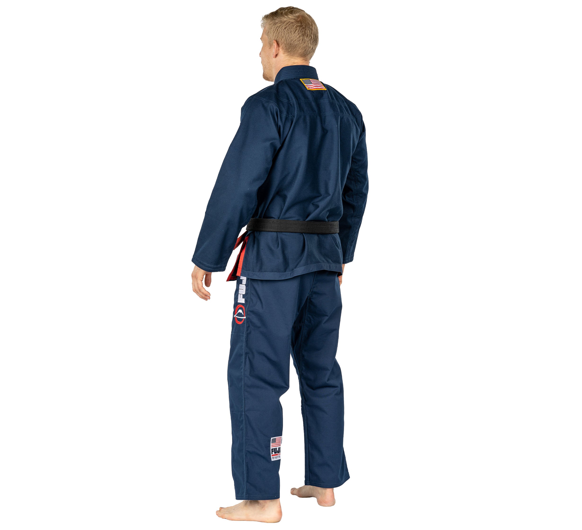 A person stands barefoot on a plain white background, facing away from the camera, wearing the Suparaito BJJ Gi 1776 Americana Edition Navy with a black belt. They have short blonde hair and martial arts patches on their gi.