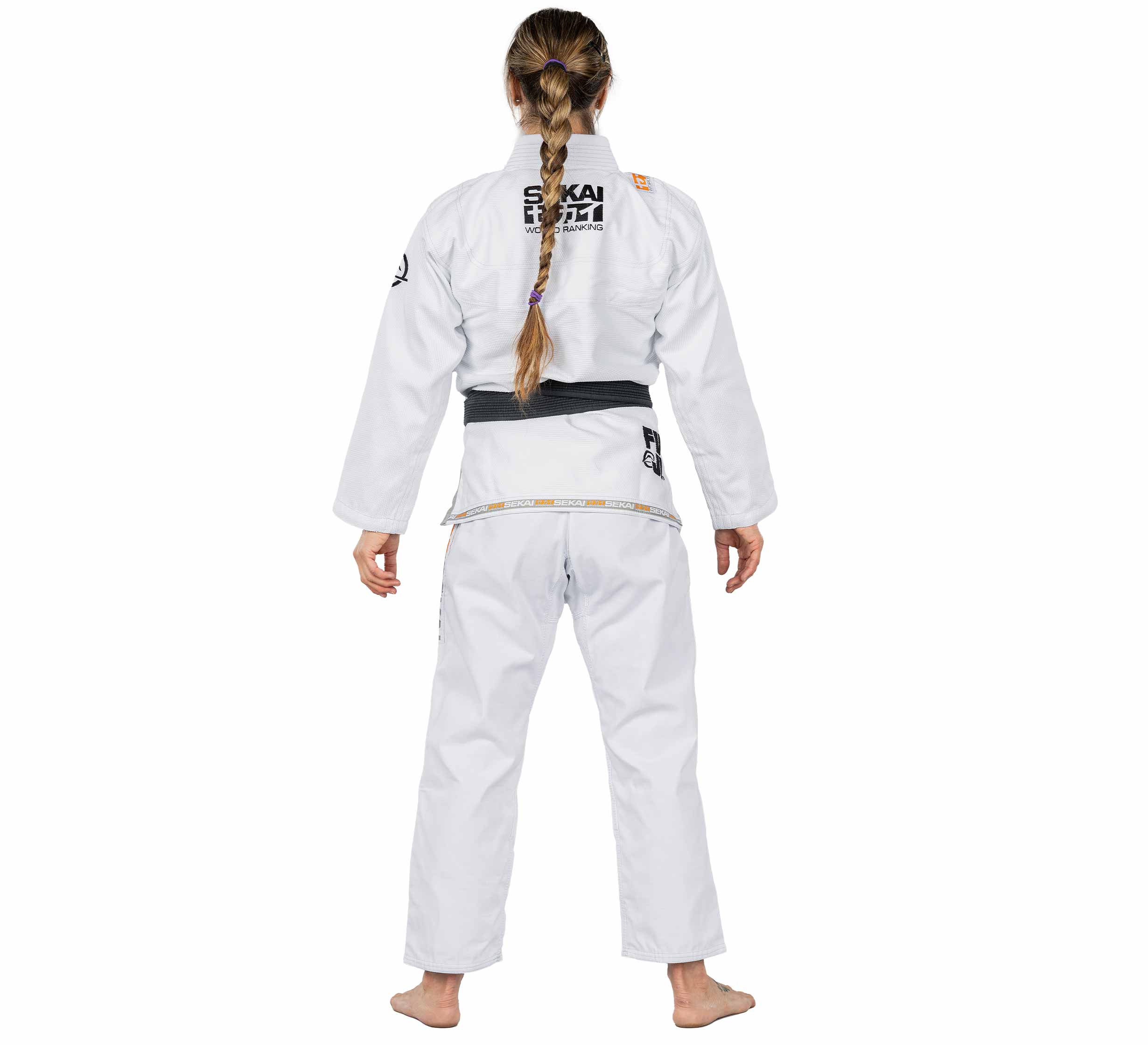 A woman with long braided hair stands barefoot, facing away from the camera, wearing the Sekai 2.0 Womens BJJ Gi White with a black belt and black-and-yellow patches on the back, arms, and waist.