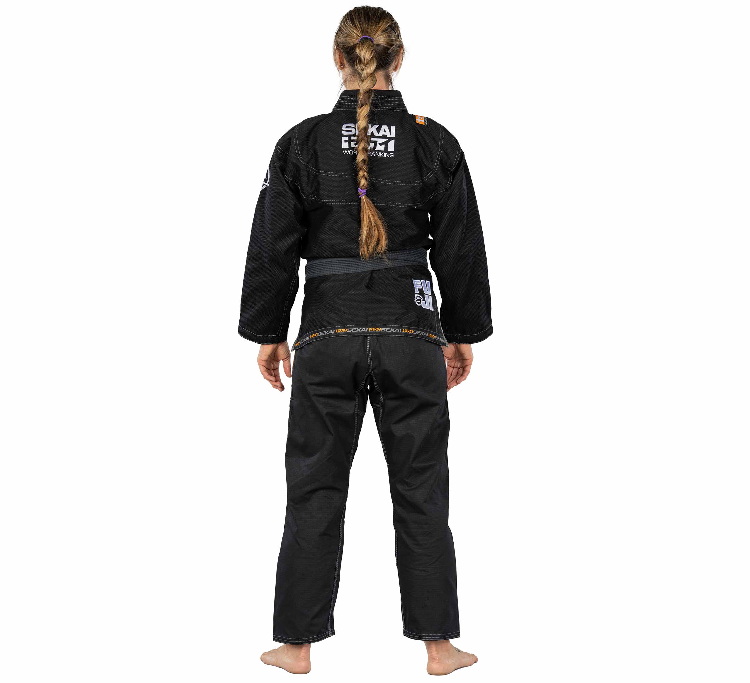A woman with long braided hair stands barefoot, facing away in a Sekai 2.0 Womens BJJ Gi Black against a white background.