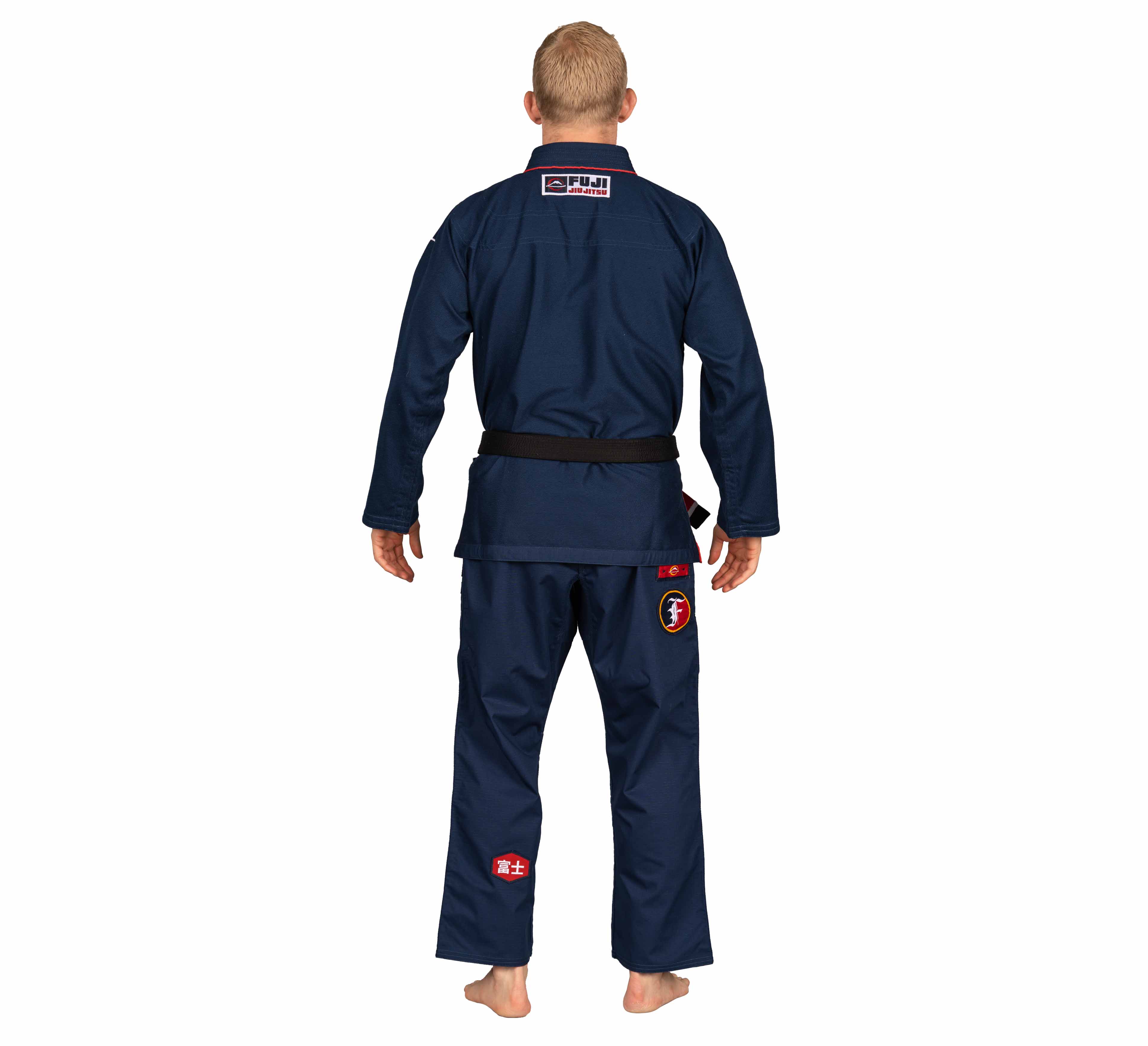 A person stands barefoot facing away, wearing the Suparaito BJJ Gi XTR Edition Navy with a black belt and displaying patches on the back, pants, and hip.