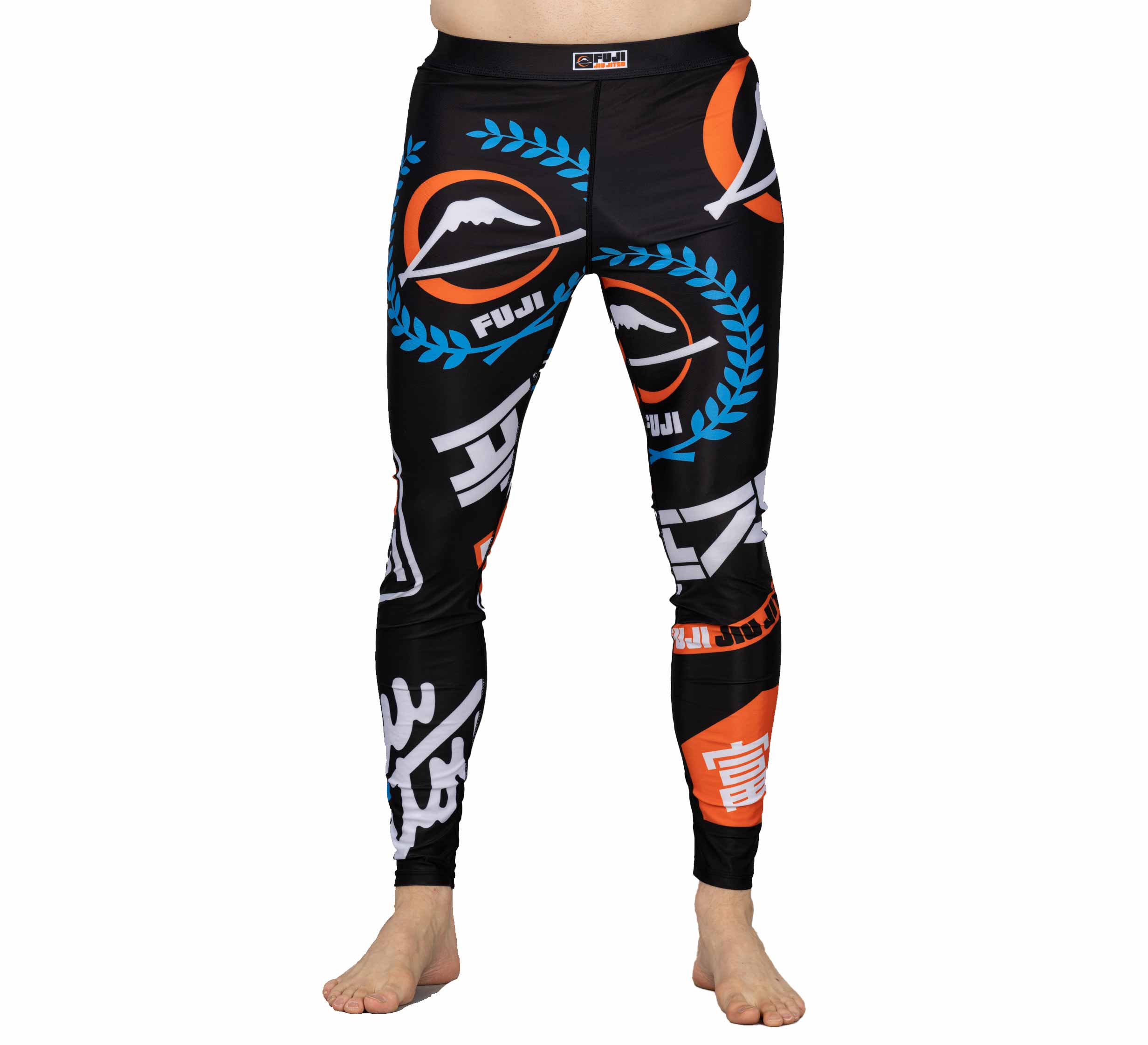 A person stands barefoot wearing XTR Extreme V1 Grappling Spats Black, featuring colorful logos with mountain graphics, laurel wreaths, and blue, orange, and white Japanese-style characters. The upper body is not visible.