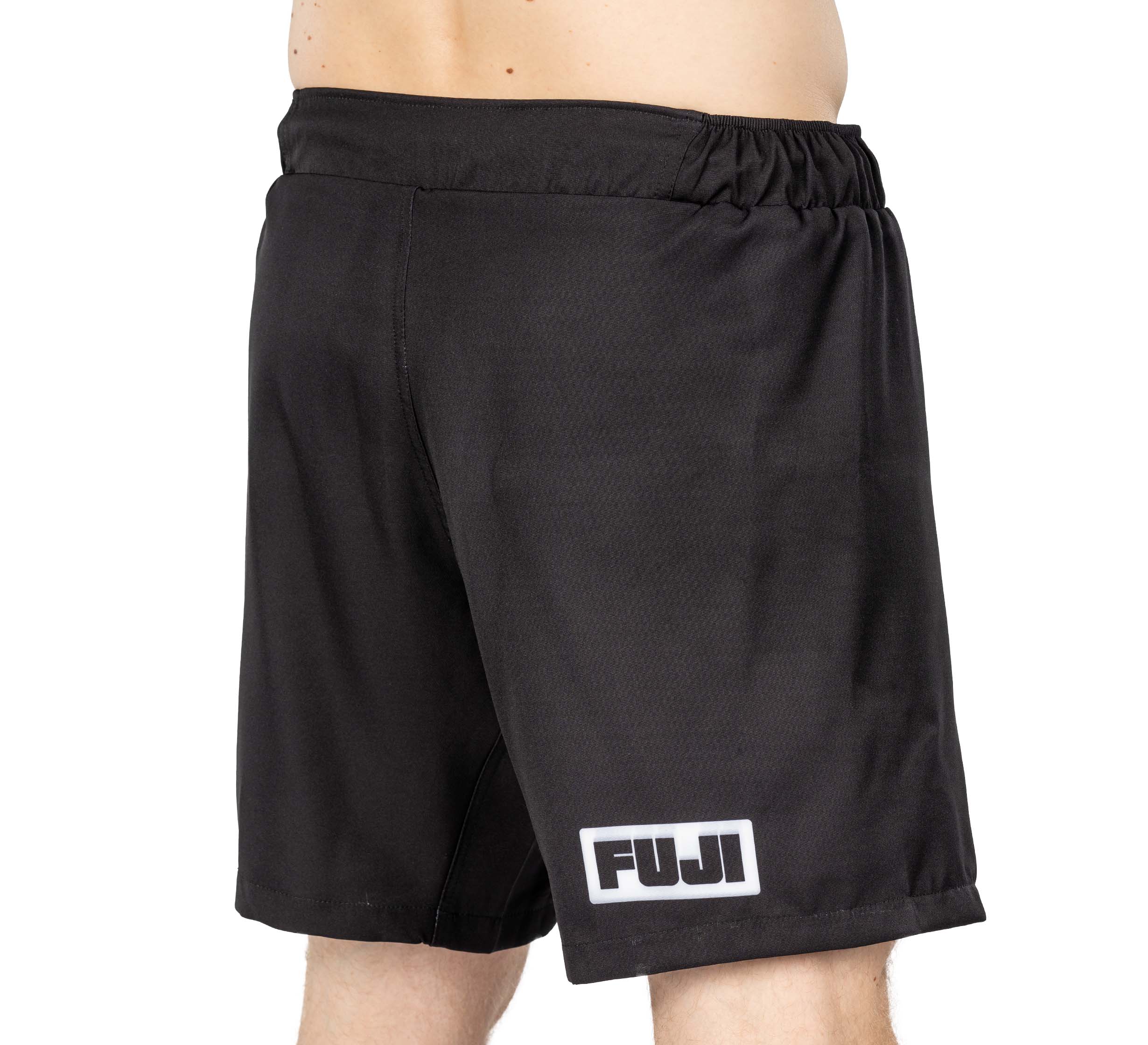 A person wearing Ultimate Grappling Shorts Black featuring an elastic waistband and a white rectangular FUJI logo on the lower left leg. Only the lower torso and upper legs are visible.