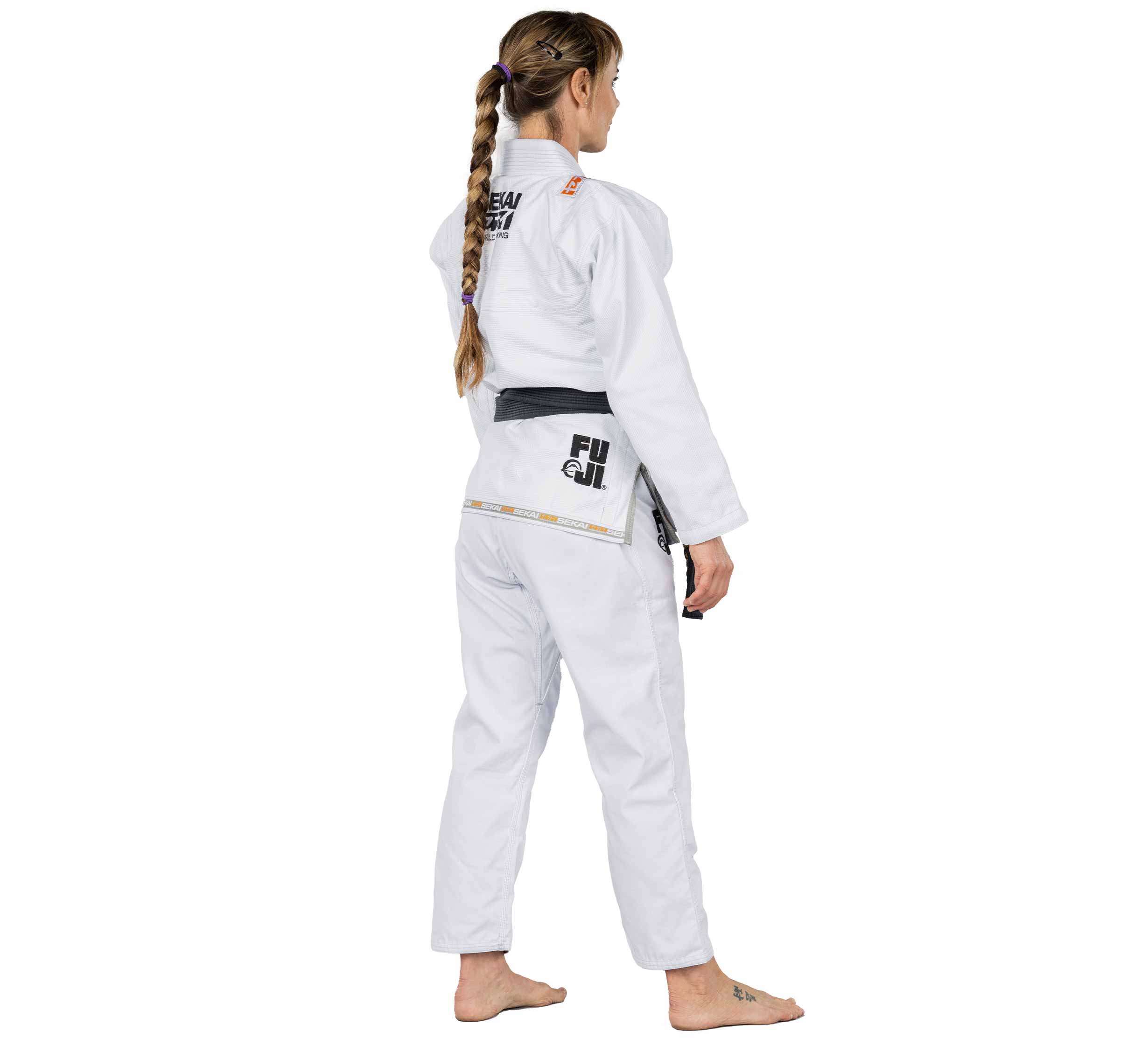 A barefoot woman with a long braid faces away, wearing the Sekai 2.0 Womens BJJ Gi White and a black belt, with Fuji branding visible on the uniform.