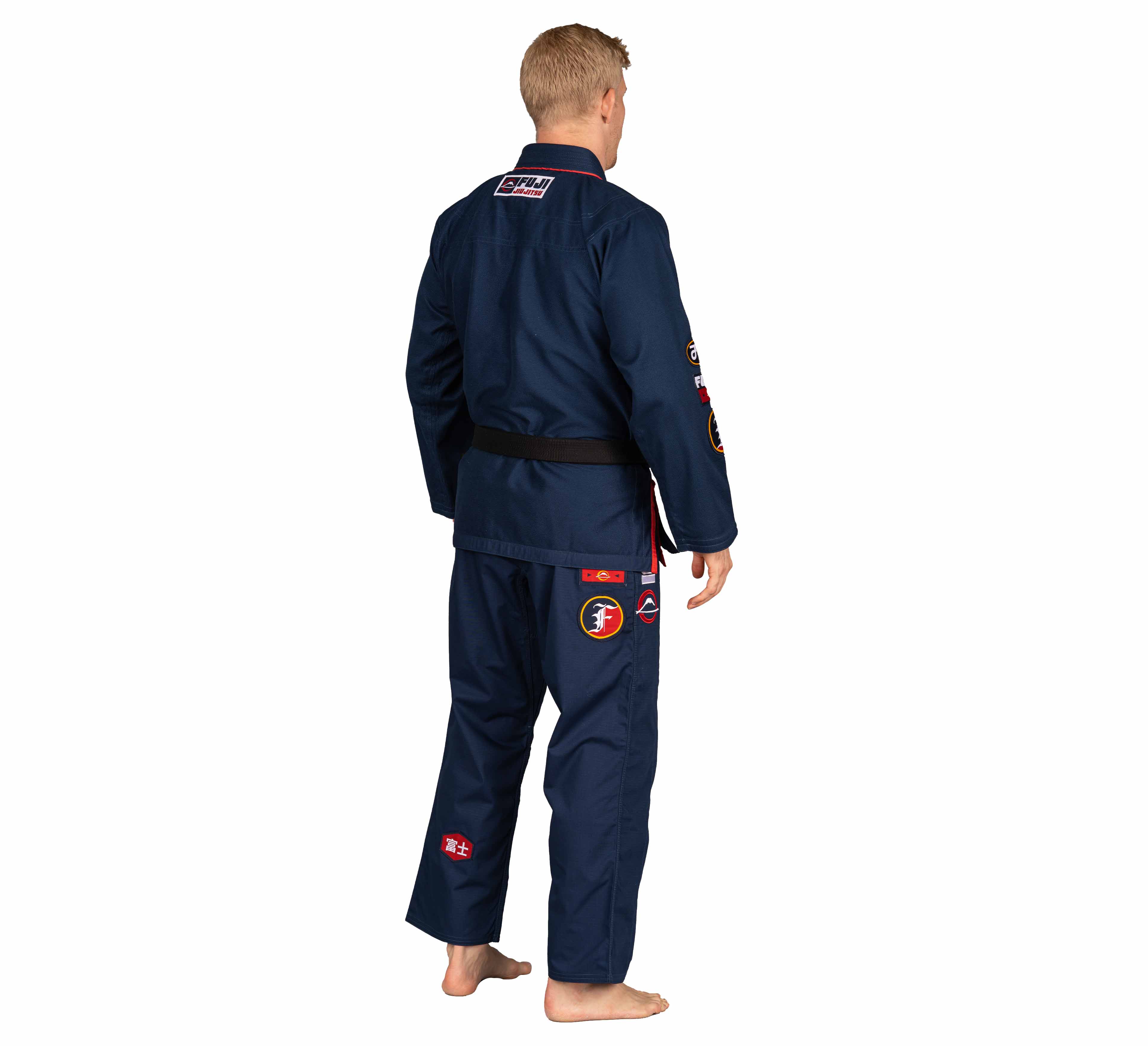 A barefoot, blonde-haired person faces away, wearing a Suparaito BJJ Gi XTR Edition Navy with colorful patches and a black belt, set against a plain white background.