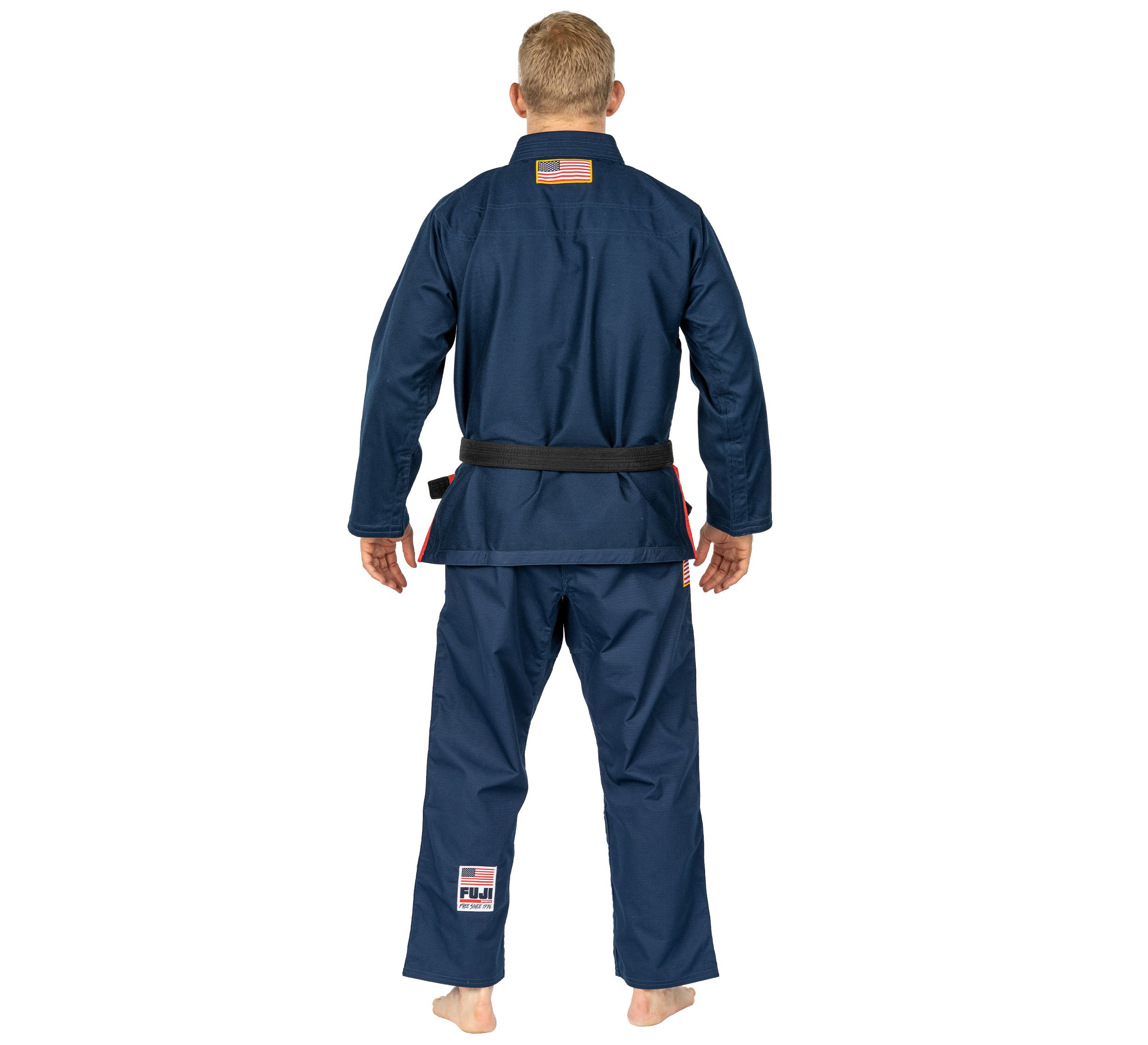 A man stands barefoot facing away, wearing the Suparaito BJJ Gi 1776 Americana Edition Navy with a black belt and an American flag patch on the back.