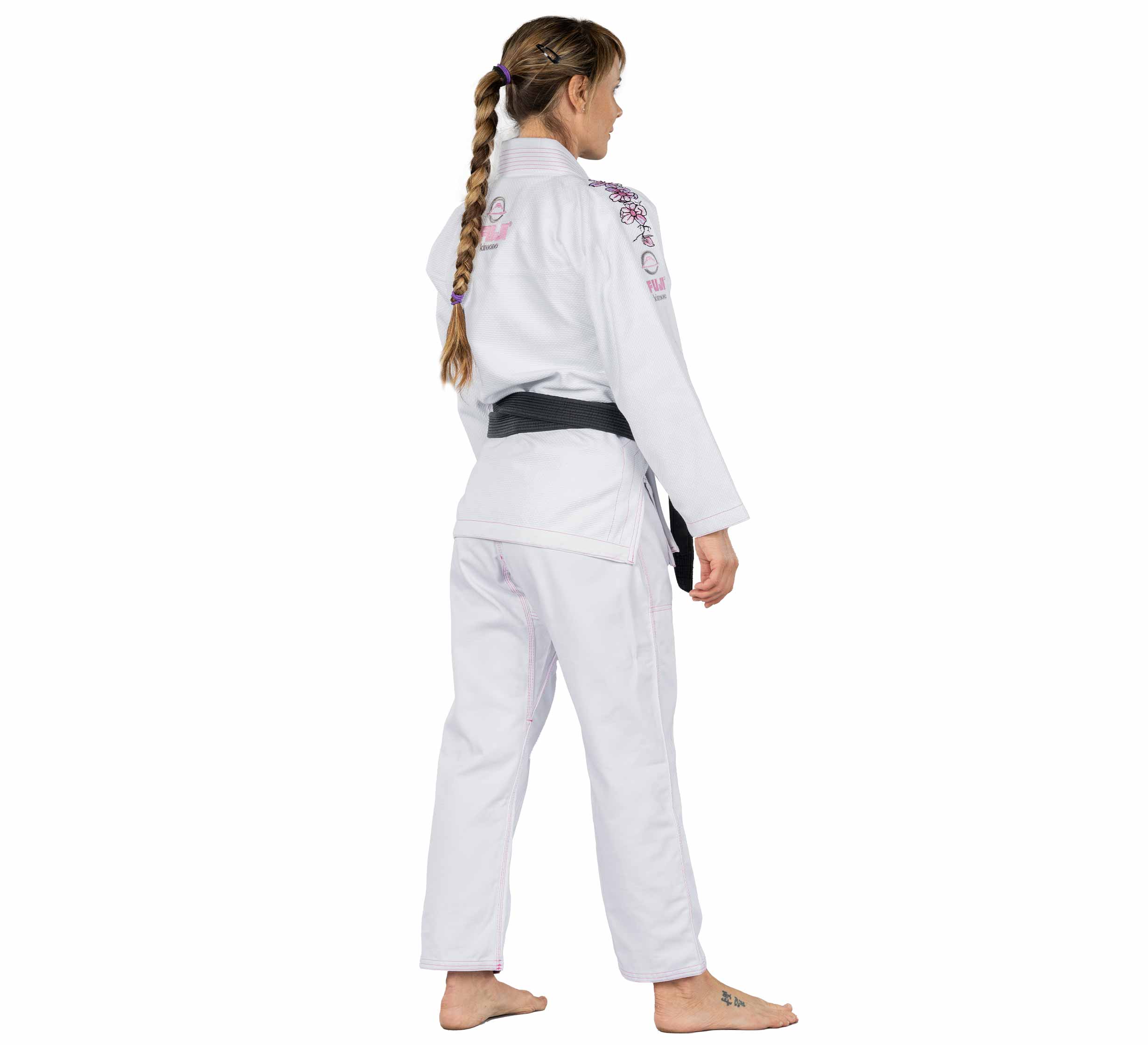 A woman stands barefoot, facing away with her head slightly turned, wearing the Blossom Womens Jiu Jitsu Gi White and a black belt, her hair styled in a braid.
