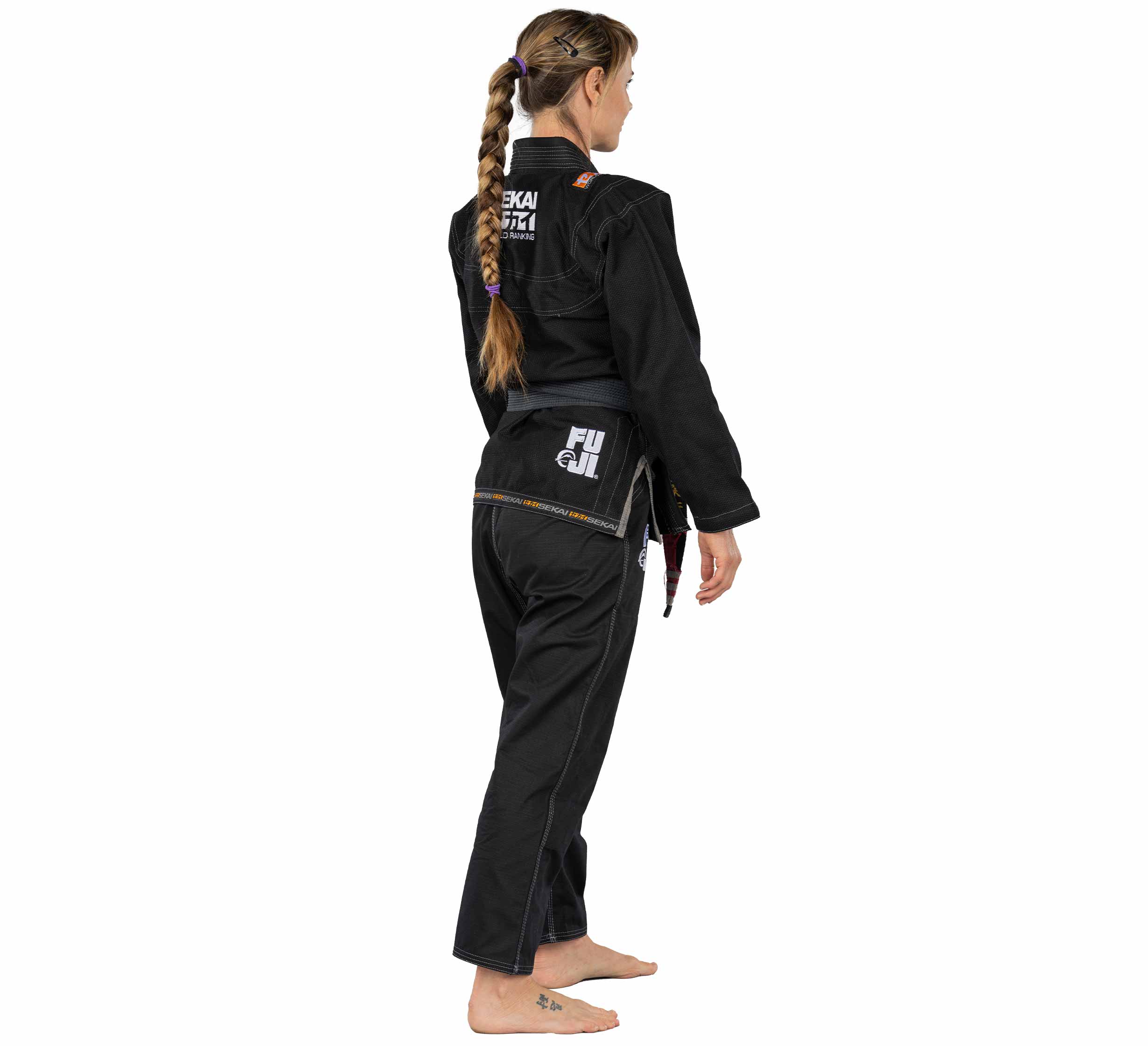 A barefoot woman with a long braid models the Sekai 2.0 Womens BJJ Gi Black, featuring patches and text, seen from the side and back against a white background.