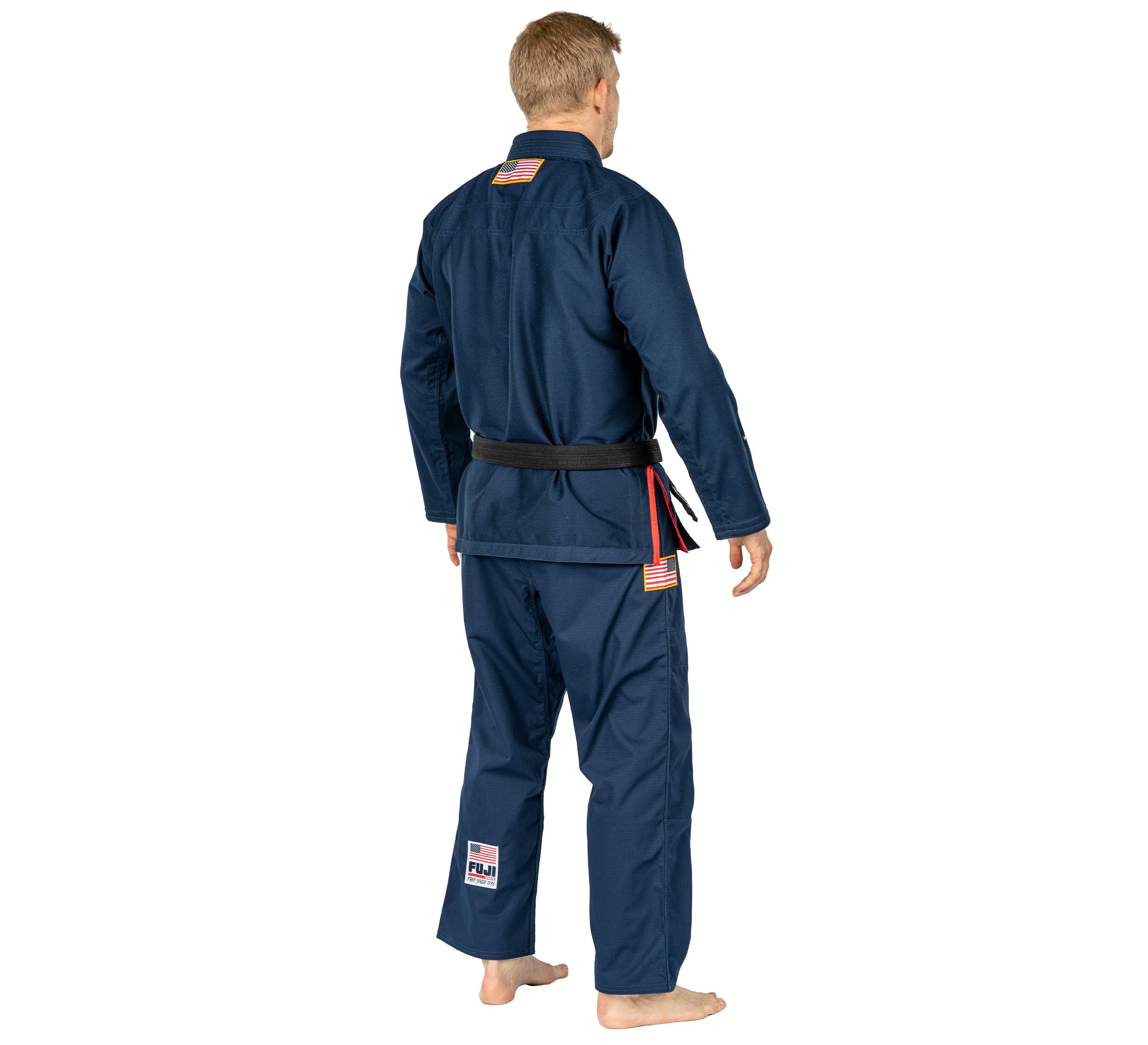 A person with short blond hair, seen from behind, wears the Suparaito BJJ Gi 1776 Americana Edition Navy with a black belt and American flag patches, standing barefoot on a white background.