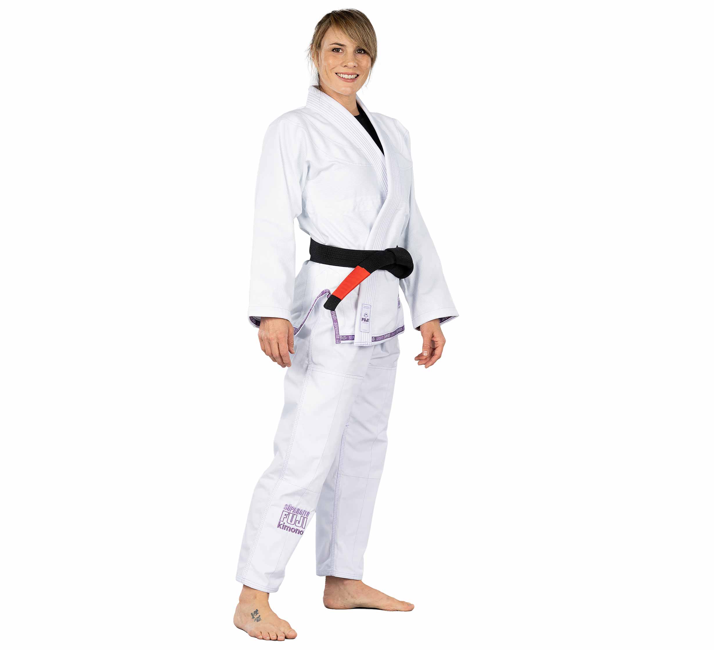 A woman stands barefoot, smiling and facing forward with relaxed arms, wearing the Suparaito Womens BJJ Gi in purple with a black belt.