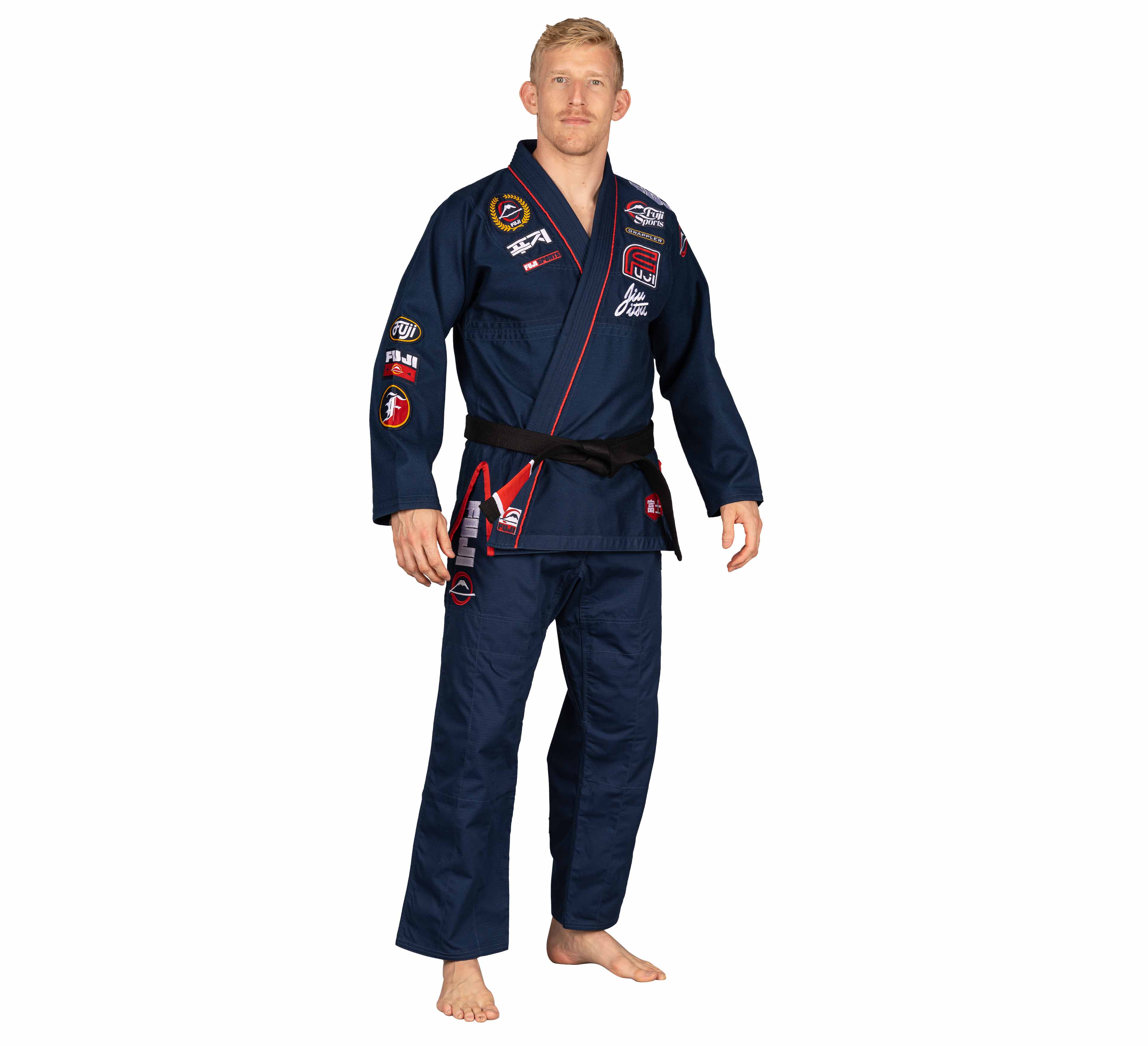 A person stands barefoot wearing the Suparaito BJJ Gi XTR Edition Navy, decorated with colorful patches and a black belt, posing against a plain white background.