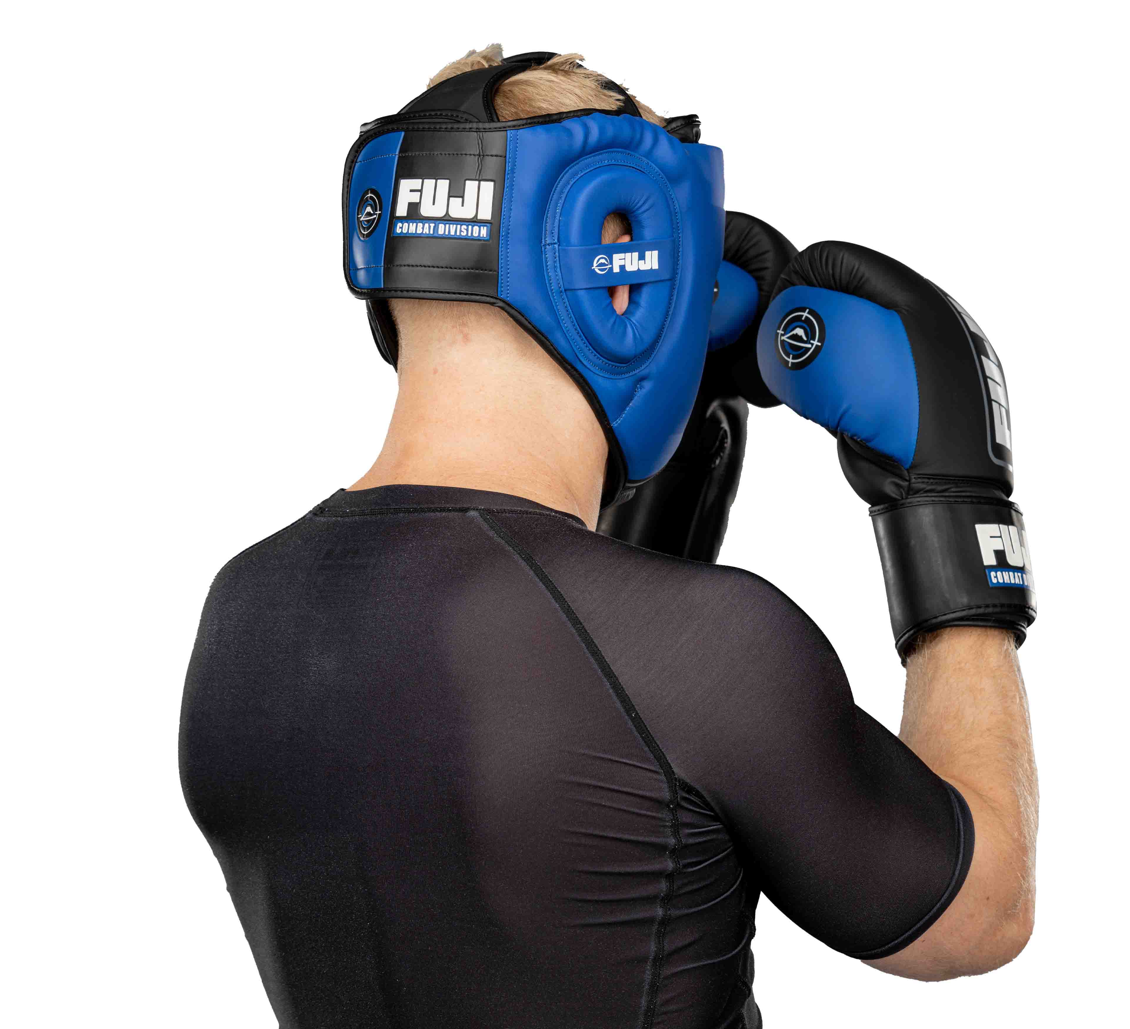 A person in a black athletic shirt and FUJI black boxing gloves stands in a defensive stance, wearing blue Precision Striking Headgear by FUJI, facing away from the camera.