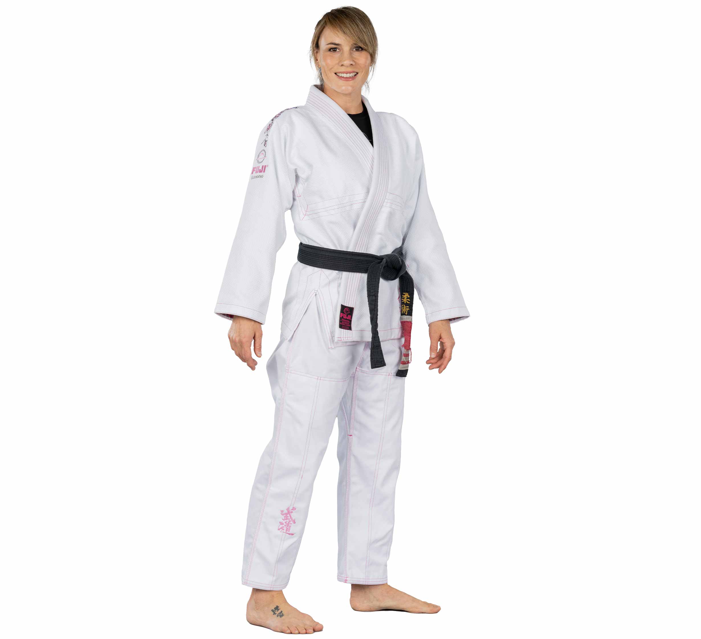 A person smiles barefoot against a plain white background, wearing the Blossom Womens Jiu Jitsu Gi in white with pink stitching and a black belt.