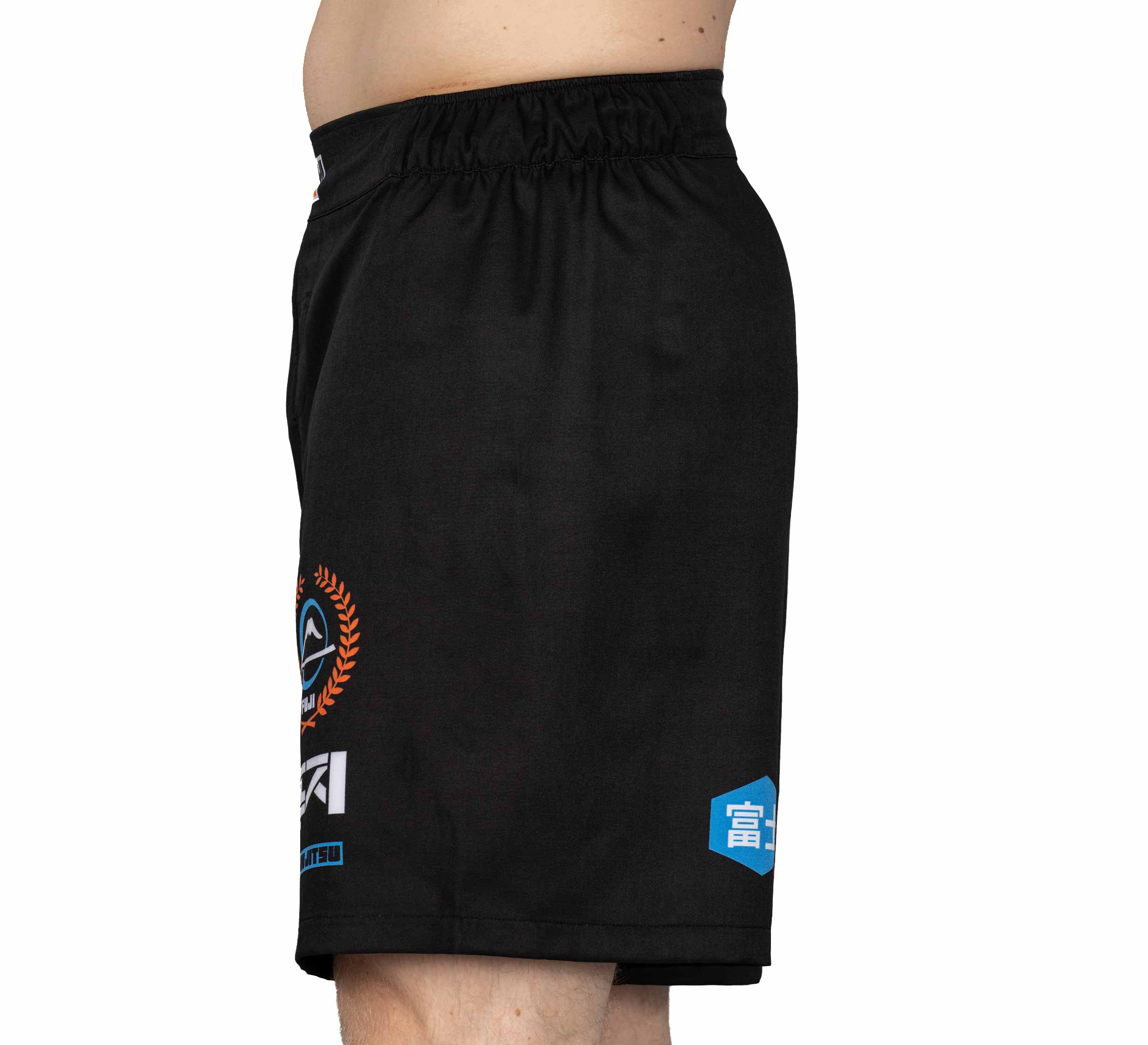 A person is wearing XTR Extreme Grappling Fight Shorts Black, featuring a stretchy waistband and multiple small logos, while standing in side profile against a plain white background.