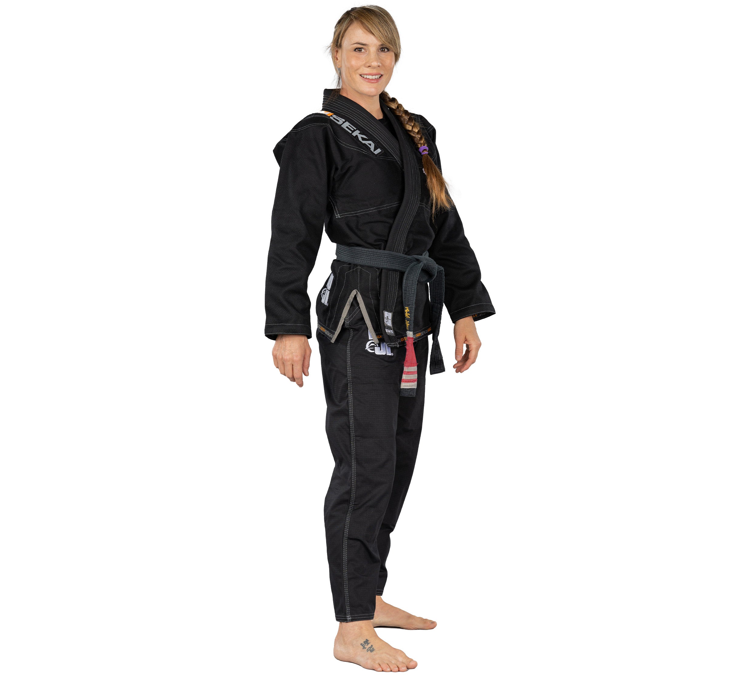 A woman with a side ponytail stands barefoot, smiling and facing slightly to the side, wearing the Sekai 2.0 Womens BJJ Gi Black with a black belt against a plain white background.