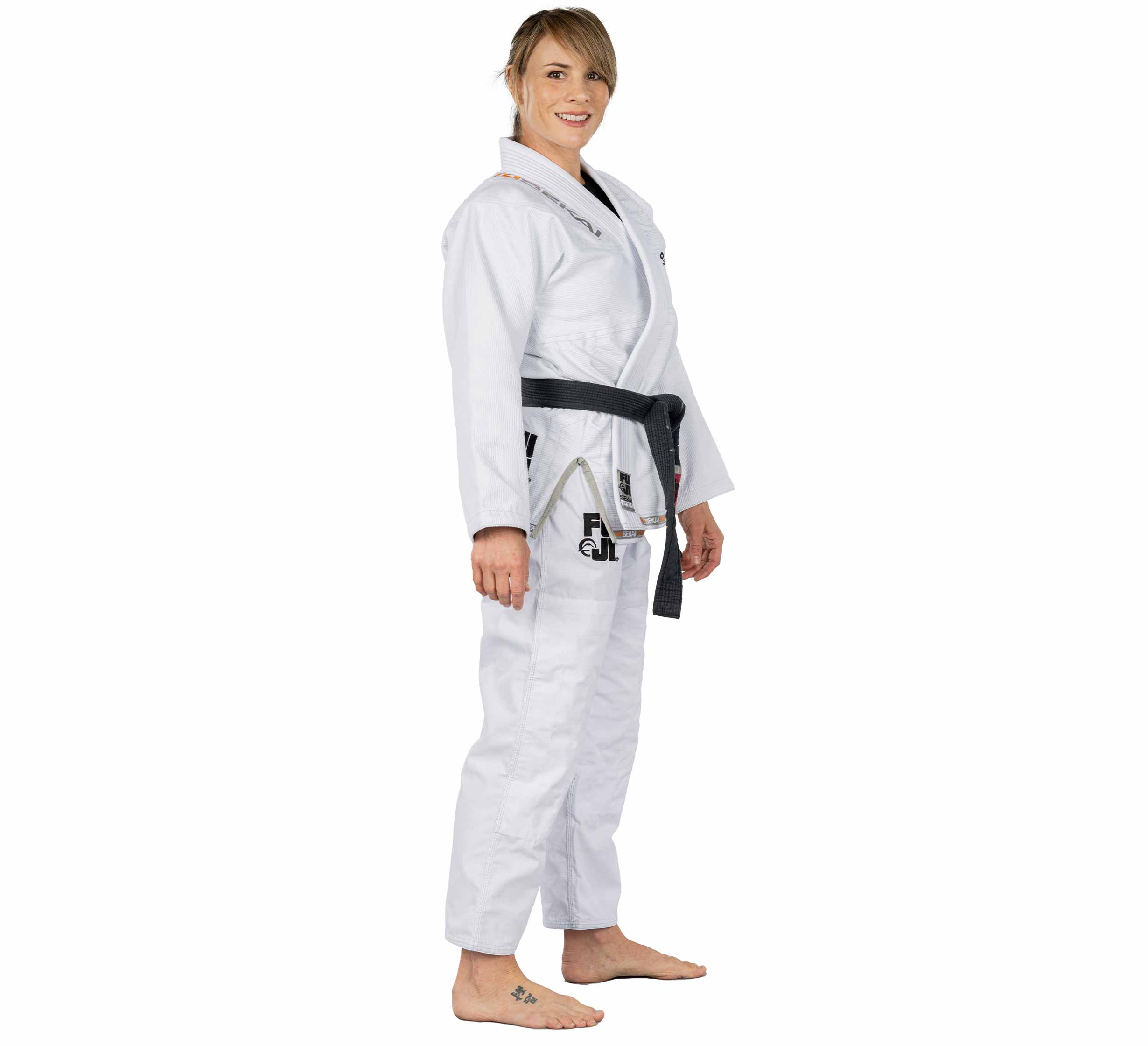 A woman wears the Sekai 2.0 Womens BJJ Gi White with a black belt, standing barefoot and smiling slightly to the side against a plain white background.