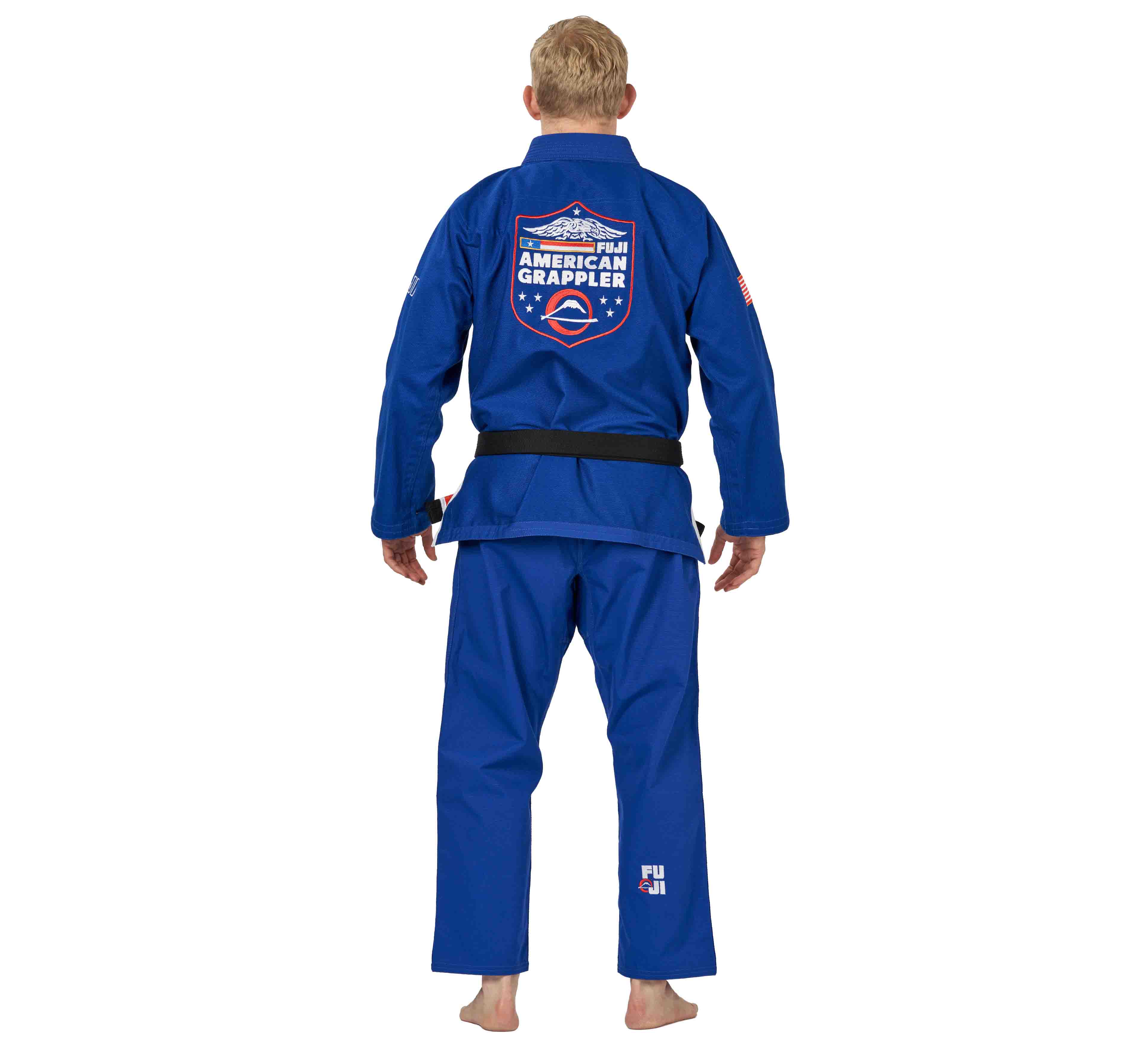 A person wearing the Suparaito BJJ Gi American Grappler Blue with a black belt stands barefoot, facing away. The gi back features a large American Grappler patch with US flag elements, and FUJI is printed near the left ankle.