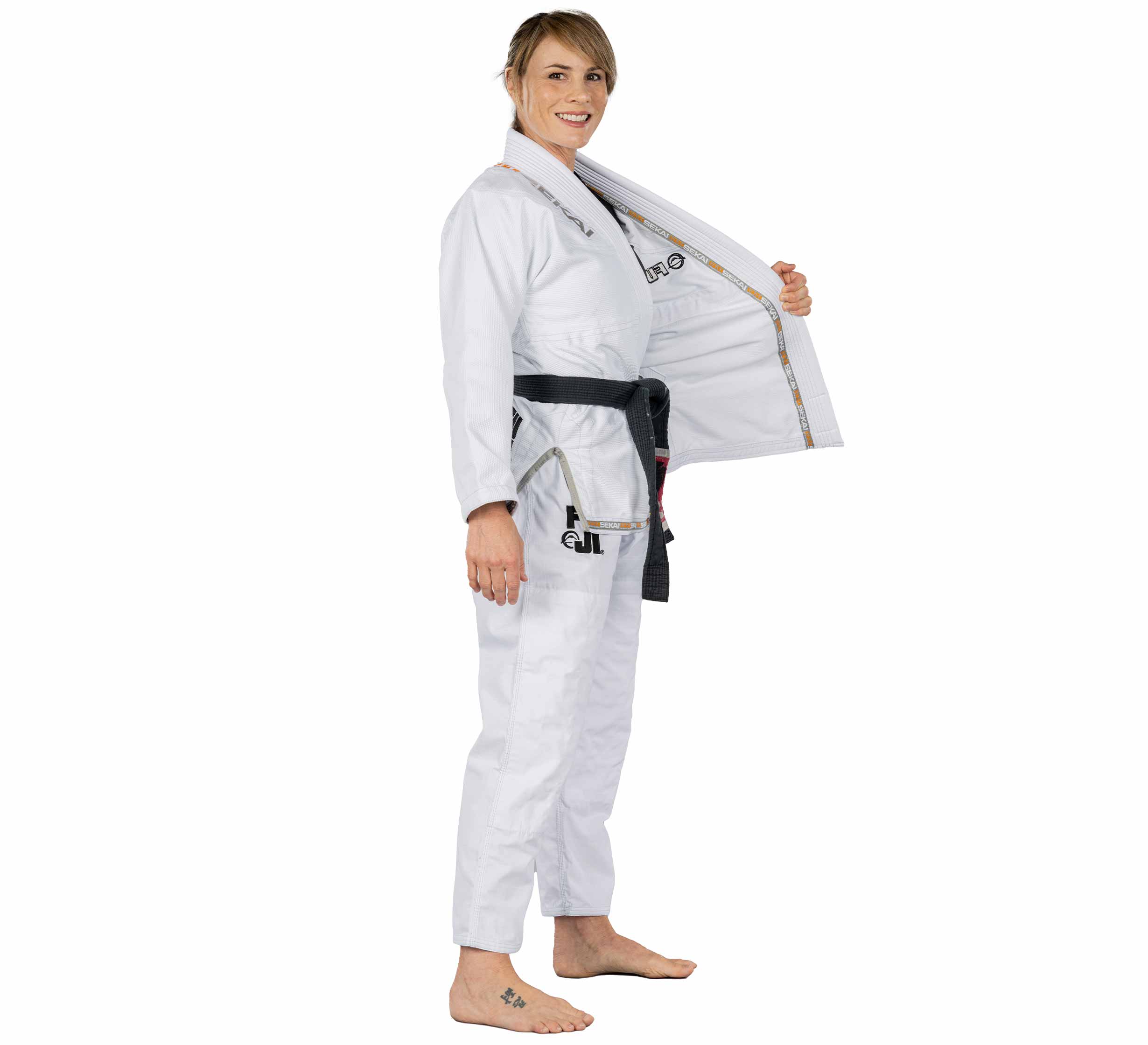 A woman smiles barefoot, wearing the Sekai 2.0 Womens BJJ Gi White with a black belt, holding one side open to show the inner lining.