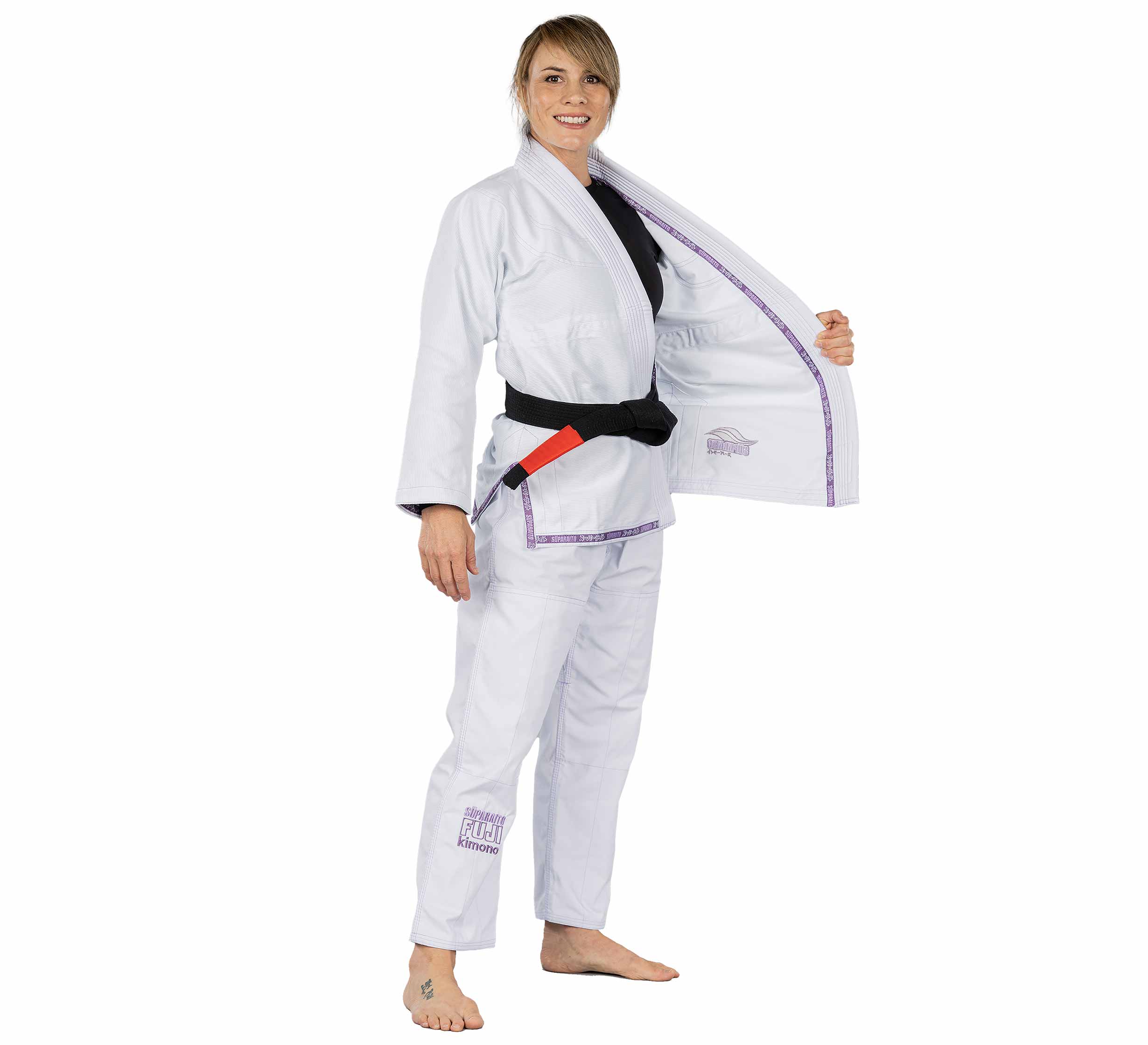 A woman wears the Suparaito Womens BJJ Gi Purple and a black belt, stands barefoot, smiles, and holds one side of her open jacket to the side.