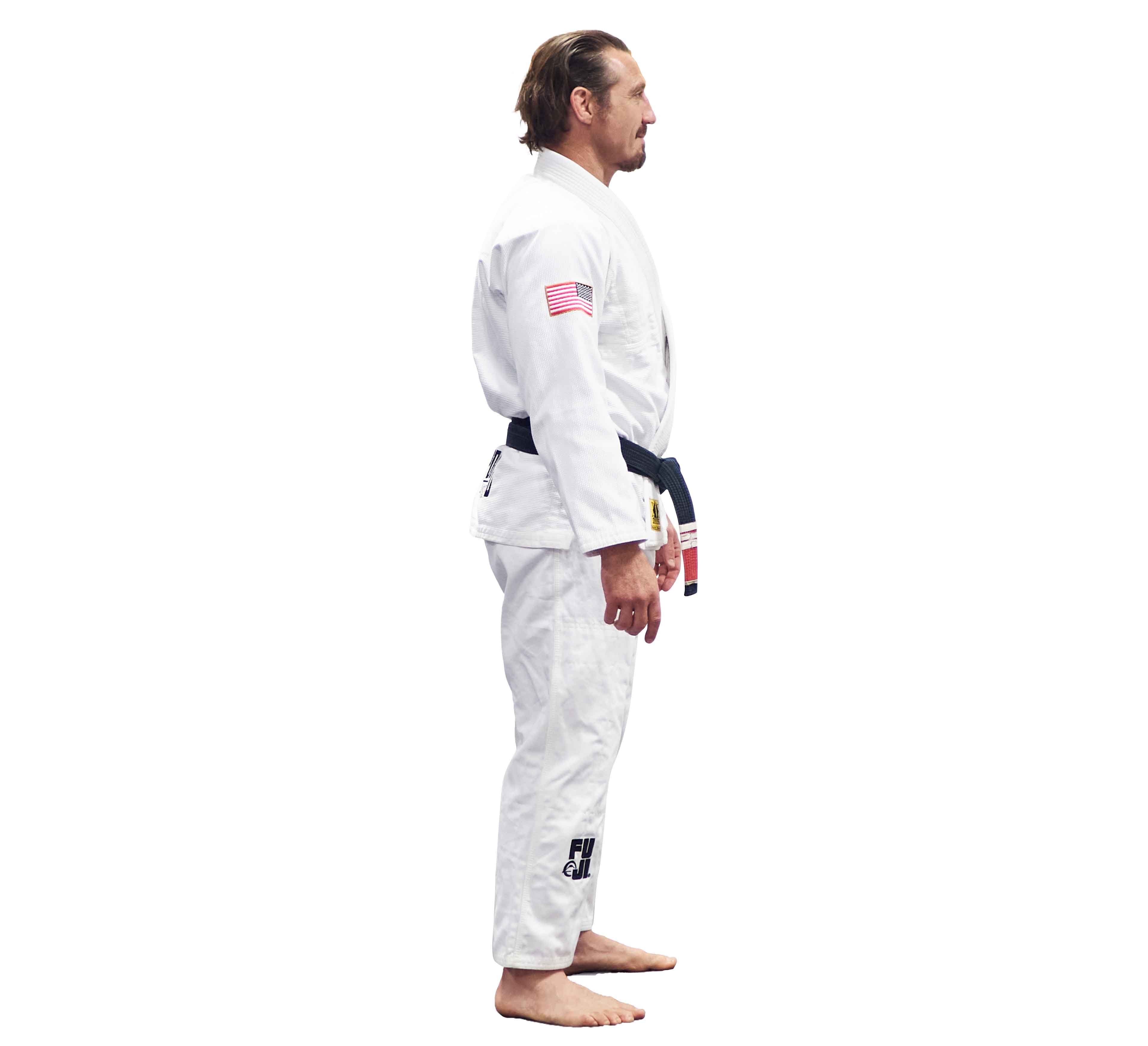 A man in the Lightweight BJJ Gi Sheepdog Response Edition White, featuring an American flag patch on his upper arm and a logo on his pants, stands barefoot in profile against a plain white background.
