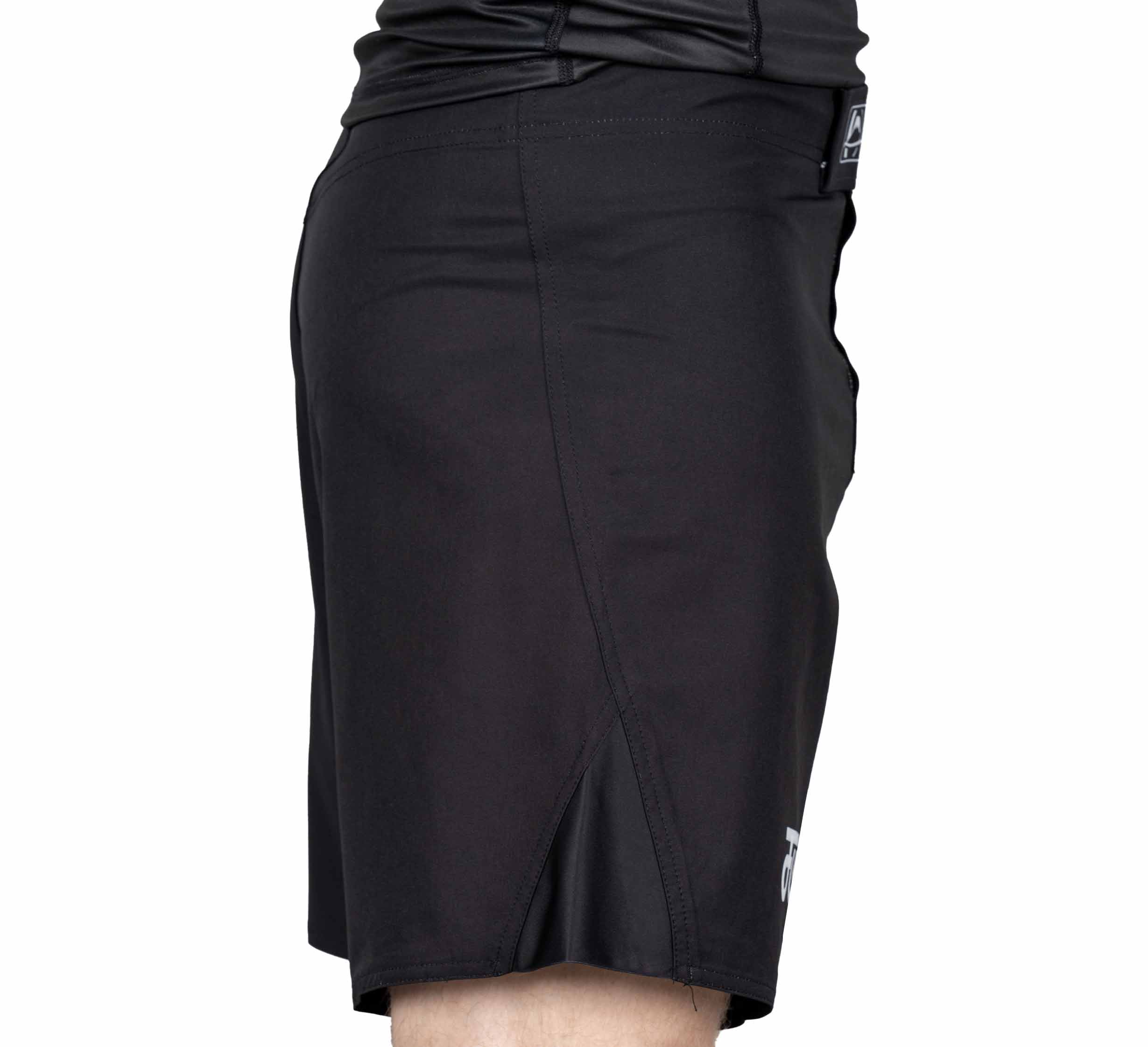 A person wearing Pro-Tech Grappling Fight Shorts in black, shown from the side with part of a black shirt at the waist and a small logo near the hem.