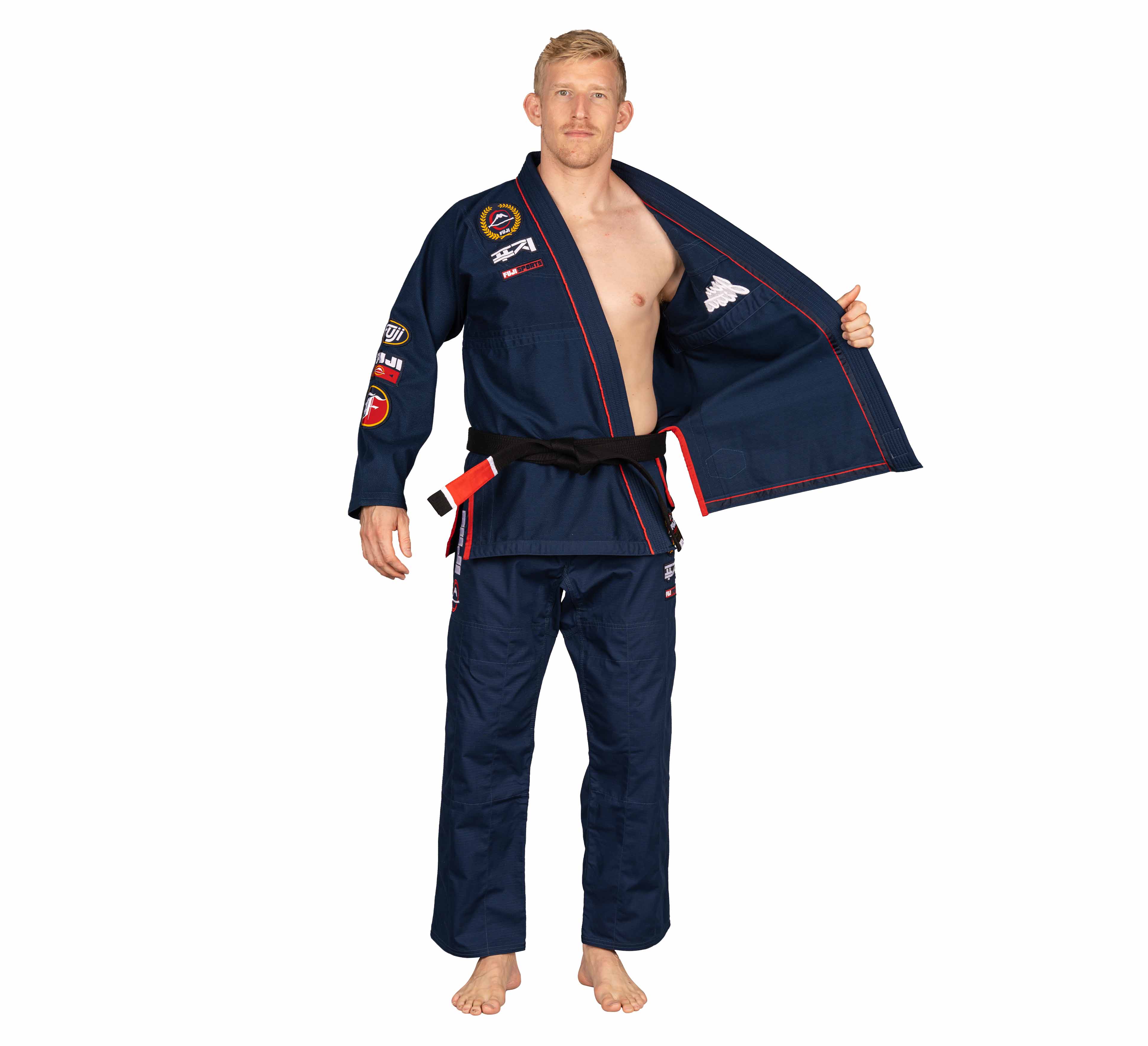 A man wearing the Suparaito BJJ Gi XTR Edition Navy with patches and a black belt stands barefoot, partially opening his gi jacket to reveal his chest against a plain white background.