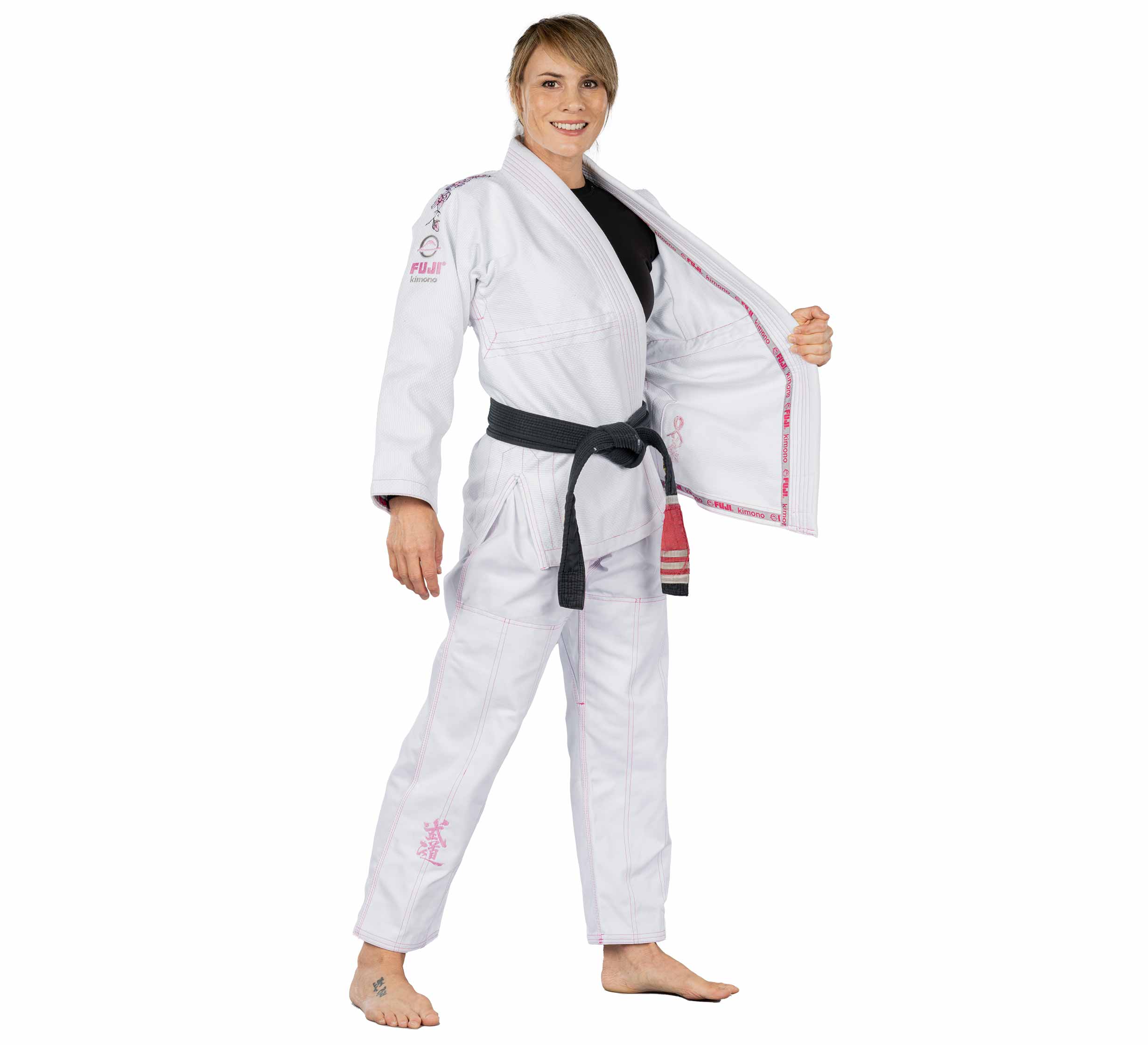 A woman wearing the Blossom Womens Jiu Jitsu Gi White with pink embroidery and brand logos stands barefoot, smiling, holding one side of her open jacket. She has a black belt and the gi features stylish detailing.