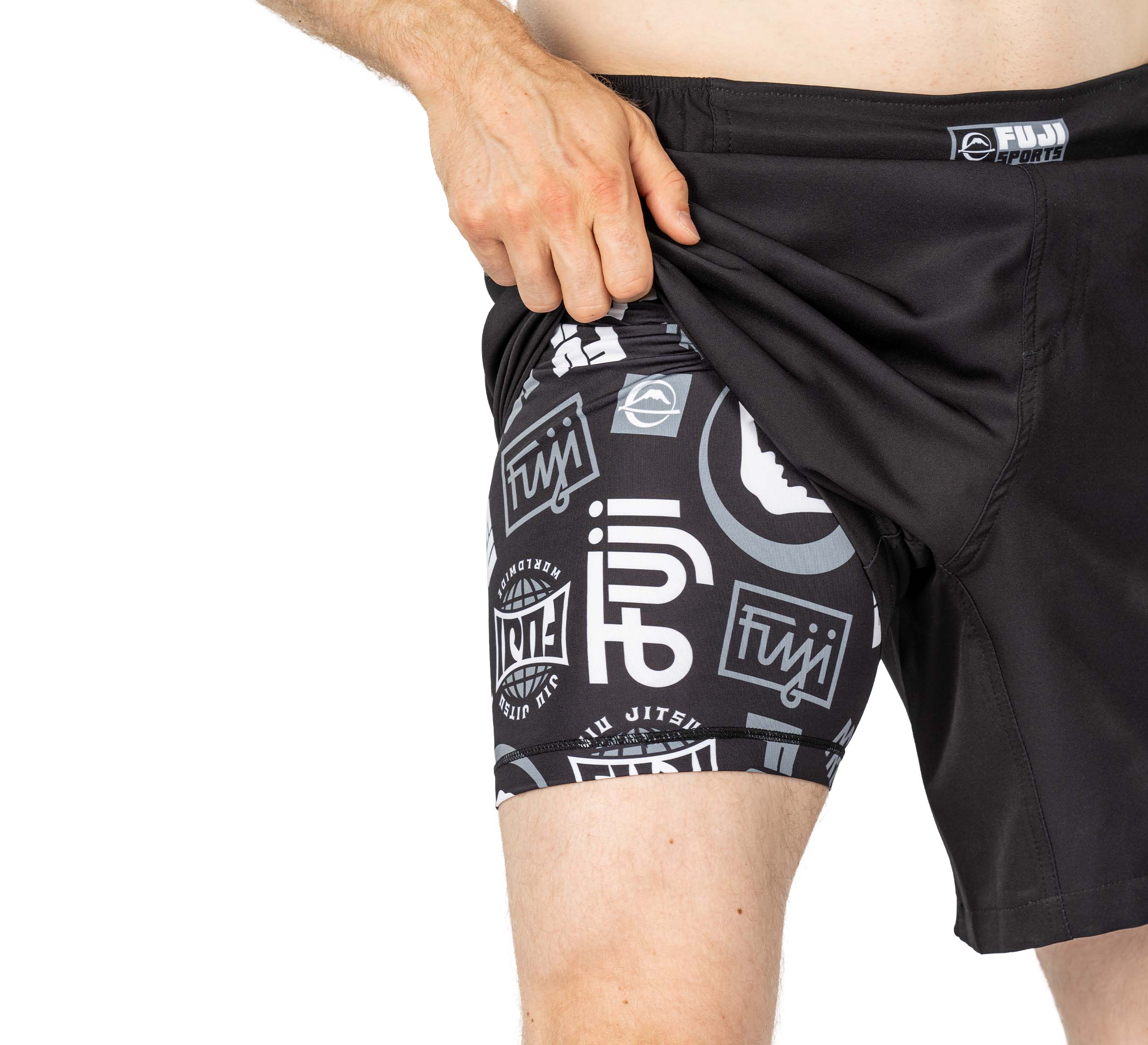 Someone lifts the outer layer of Ultimate Grappling Shorts Black to reveal matching black compression shorts underneath, both adorned with white jiu jitsu logos and patterns.