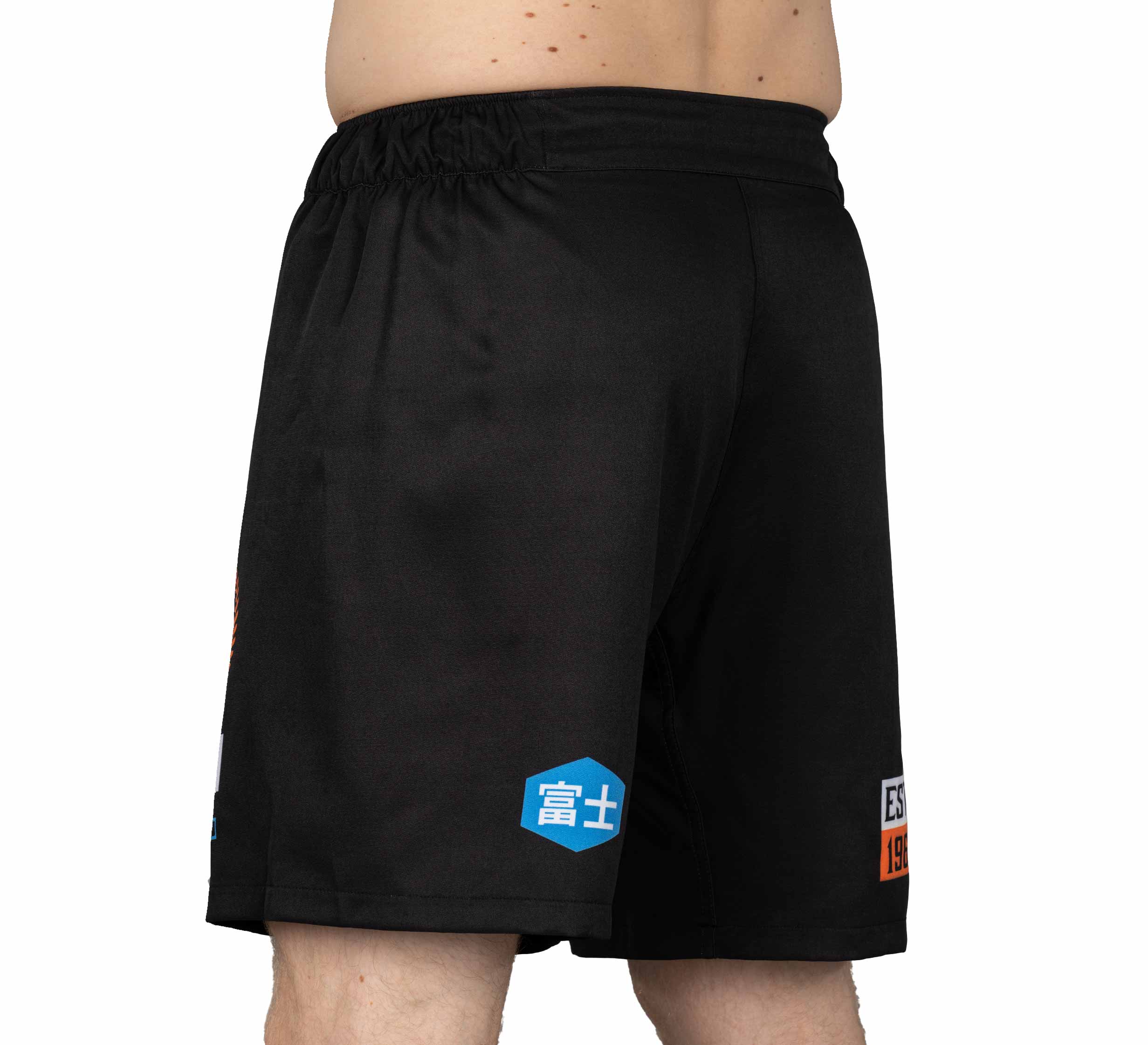 A person shown from the back, waist down, wearing XTR Extreme Grappling Fight Shorts Black with an elastic waistband, blue and white patches, and orange lettering on the left leg.