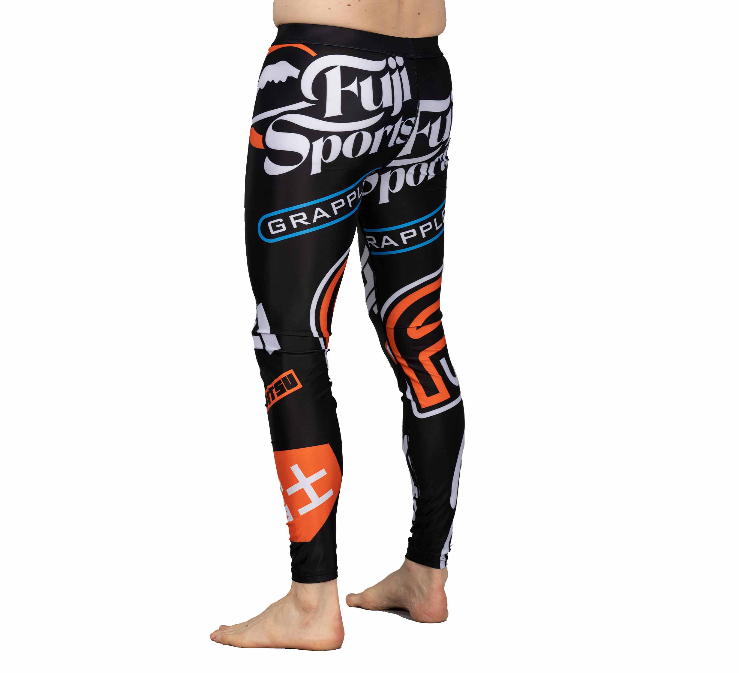 A person stands barefoot, wearing the black XTR Extreme V1 Grappling Spats by Fuji Sports with bold logos and graphic designs, photographed from the side against a white background.